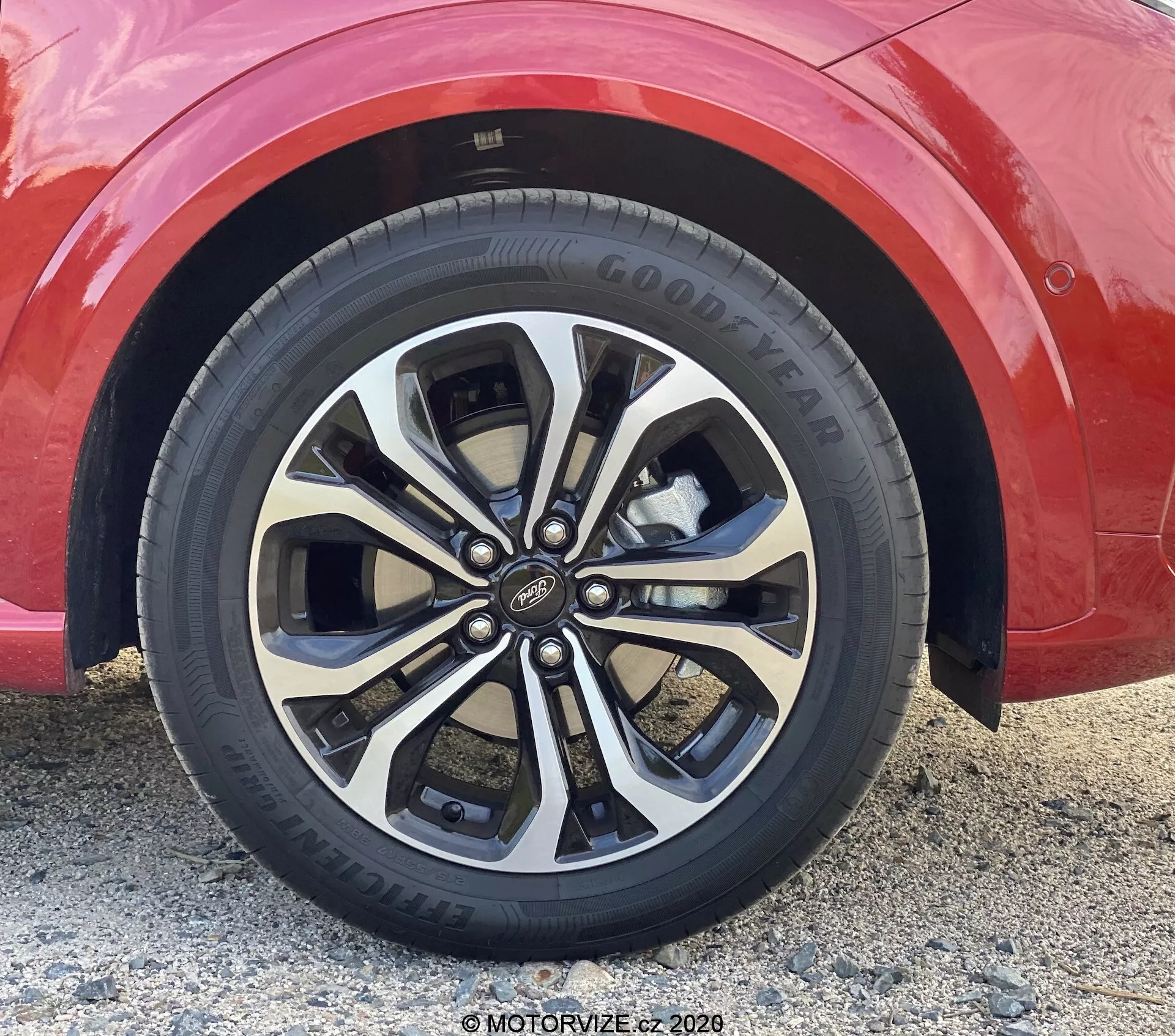 Primo piano di una ruota in lega multirazze su un'auto rossa, vista da un'angolazione bassa e leggermente inclinata verso l'alto. La ruota presenta una finitura bicolore argento e scura, con razze sdoppiate accoppiate, superficie lucida e un coprimozzo riconoscibile. È visibile anche il pneumatico Goodyear con un battistrada sostanziale e un pattern a 5 bulloni.