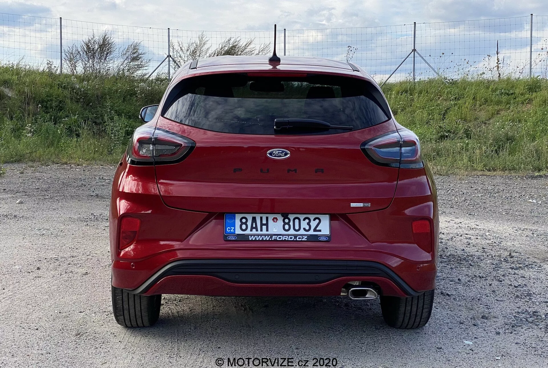 Vista posteriore di una Ford Puma rossa del 2020 hatchback su una superficie di ghiaia, catturata da un'angolazione livellata e centrata. Le caratteristiche includono uno spoiler posteriore, tergicristallo posteriore, luce di stop alta, sistema di scarico doppio, superfici riflettenti, sensori di parcheggio, telecamera posteriore ed emblema Ford e nome modello in evidenza. Nessun attrezzo di traino o portapacchi visibile. Foto di MOTORVIZE.cz.