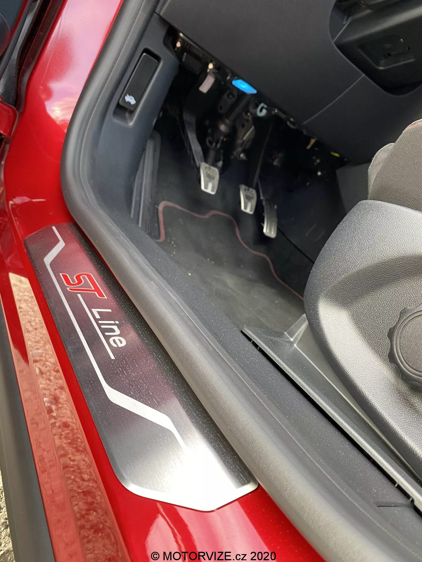 Vista ravvicinata della soglia della porta di un veicolo Audi con il badge 'S Line' in argento e rosso su sfondo nero. La ripresa dall'alto e leggermente verso l'interno cattura il bordo inferiore della porta, il bordo interno e una porzione del vano piedi del conducente con i pedali del freno e dell'acceleratore visibili. La luce naturale del giorno illumina la scena, evidenziando il colore rosso dell'esterno e i componenti interni vicino al cruscotto.
