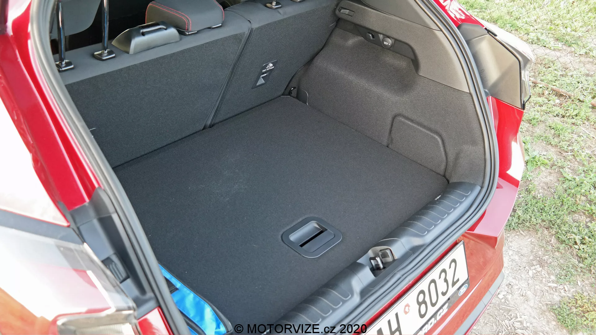 L'immagine mostra il vano posteriore o bagagliaio di una Ford Puma 2020. La foto è scattata da un angolo leggermente in alto e di lato, fornendo una vista chiara dell'interno spazioso del bagagliaio e della sua soglia. La moquette scura suggerisce che è progettata per minimizzare la comparsa di sporco. Non sono visibili carichi, sottolineando la pulizia e la spaziosità del bagagliaio. Le caratteristiche visibili includono rientranze laterali o tasche per lo stoccaggio di piccoli oggetti e un gancio o punto di ancoraggio per fissare gli oggetti. Il gruppo ottico posteriore dell'auto è parzialmente visibile sul lato destro dell'immagine.