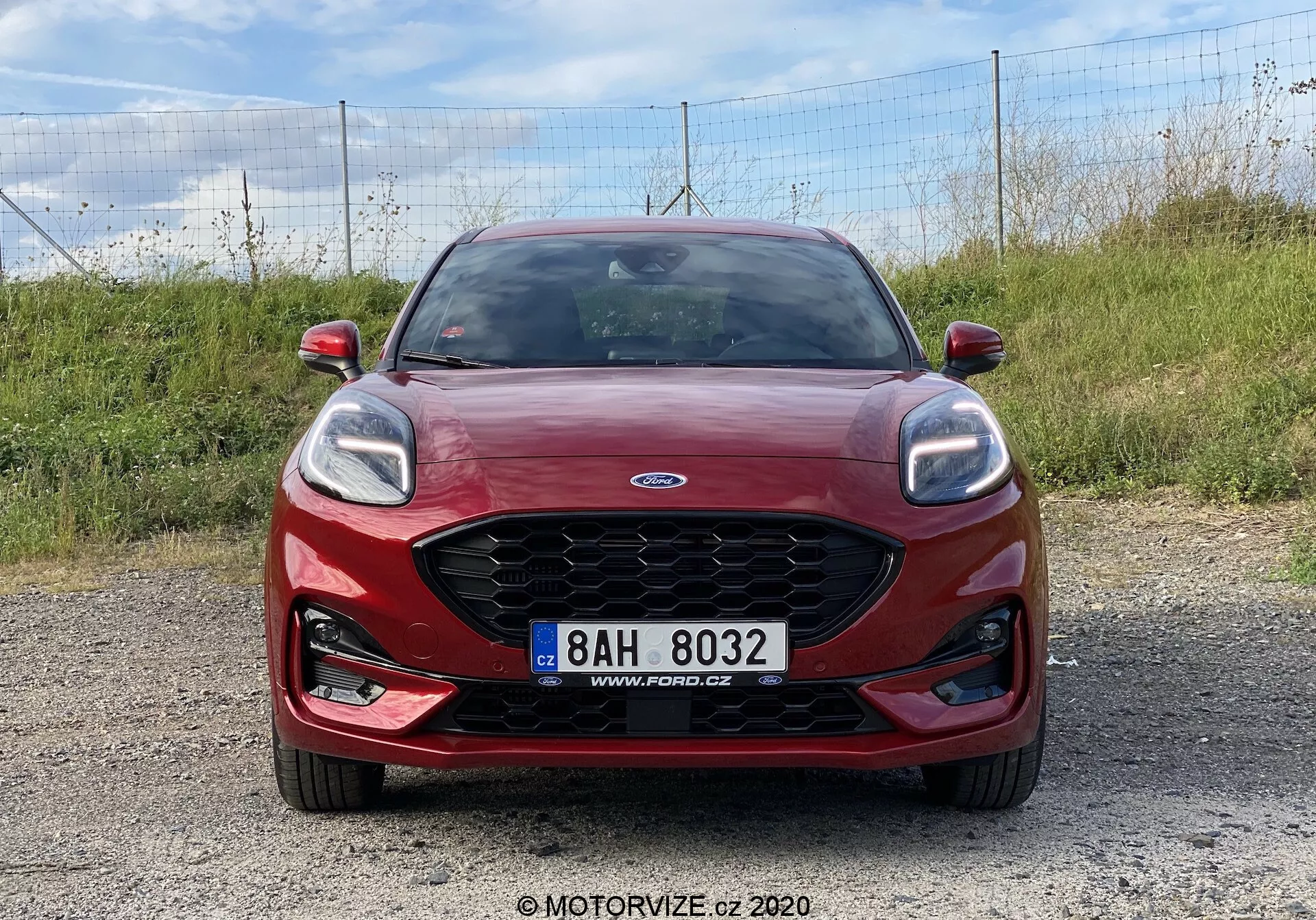 Ford Puma 2020 rosso catturato da un'angolazione frontale su una superficie di ghiaia, mostrando la distintiva griglia Ford con logo centrato, fari eleganti con luci diurne a LED, fari fendinebbia integrati e targa montata centralmente. Ideale per il design del frontale del veicolo, l'identificazione del marchio e i moderni sistemi di illuminazione automobilistica.