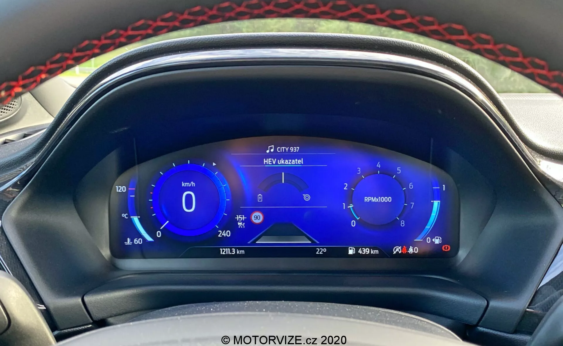Vista ravvicinata del cruscotto digitale del Ford Puma 2020 visto dal sedile del conducente. Il display digitale include un tachimetro con intervalli di velocità ecologica sulla sinistra, un contagiri al centro, indicatori di carburante ibrido a destra e ulteriori informazioni come chilometraggio, distanza del viaggio, autonomia e temperatura esterna in basso. Al centro in alto appare una notifica con la scritta 'HEV úzkazník!', che indica la modalità veicolo elettrico ibrido. Il design elegante e orientato alla tecnologia con toni di blu, bianco e rosso evidenzia i sistemi di informazione avanzati per il conducente e la tecnologia ibrida del veicolo.