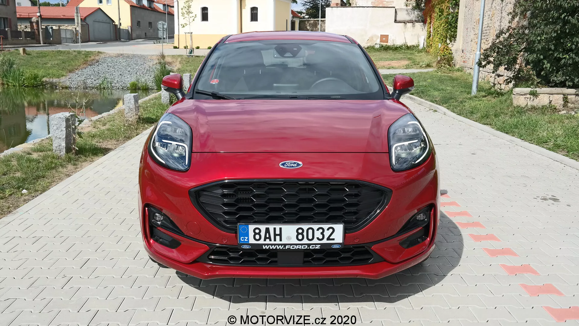 Vista frontale di una Ford Puma rossa del 2020, con griglia esagonale a rete nera, fari moderni inclinati all'indietro, paraurti anteriore sportivo con ampie prese d'aria e targa centrale. Le linee di contorno del cofano dinamico migliorano l'estetica del veicolo. Sullo sfondo, un'area residenziale.