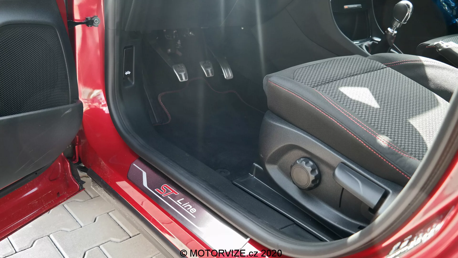 Vista ravvicinata dell'interno di una Ford Puma 2020, con particolare attenzione alla soglia della porta in metallo con marchio S Line, sedile sportivo in misto pelle e tessuto con rinforzi laterali, comandi elettronici per la regolazione del sedile, pedali in metallo con inserti in gomma per cambio manuale, tappetino del pavimento in moquette, pannello interno rosso e nero con griglia dell'altoparlante e luce solare diretta che illumina l'interno.