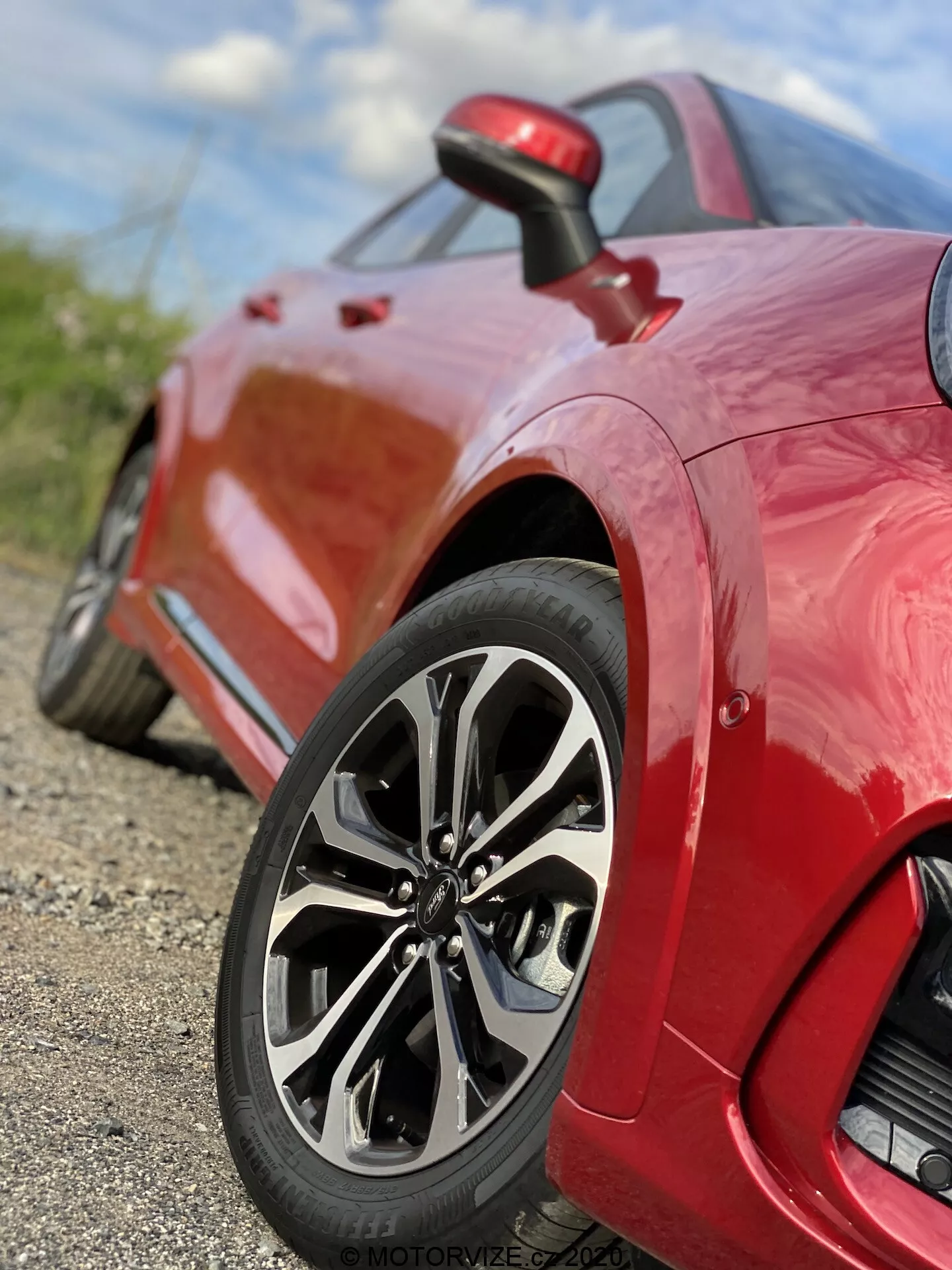 Vista ravvicinata di un'auto sportiva Ford Puma 2020 rossa, incentrata sulla ruota posteriore e il pannello laterale. Lo scatto da terra, angolato verso il basso, mette in evidenza il sofisticato design multiraggio della ruota, il pneumatico a profilo basso e la finitura lucida della vernice. L'immagine cattura dettagli intricati del design della ruota e del battistrada, con una bassa profondità di campo che mantiene la ruota a fuoco mentre lo sfondo è sfocato. Una piccola presa d'aria visibile dietro la ruota sul pannello laterale enfatizza le caratteristiche di prestazione e aerodinamiche del veicolo.