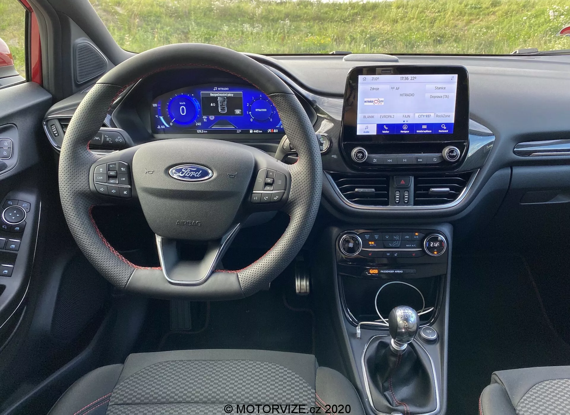 Interno della Ford Puma 2020 dalla prospettiva del guidatore, che mostra il volante multifunzionale a tre razze, il quadro strumenti digitale, la console centrale di infotainment con funzioni di connettività, il cambio automatico e vari sistemi di controllo