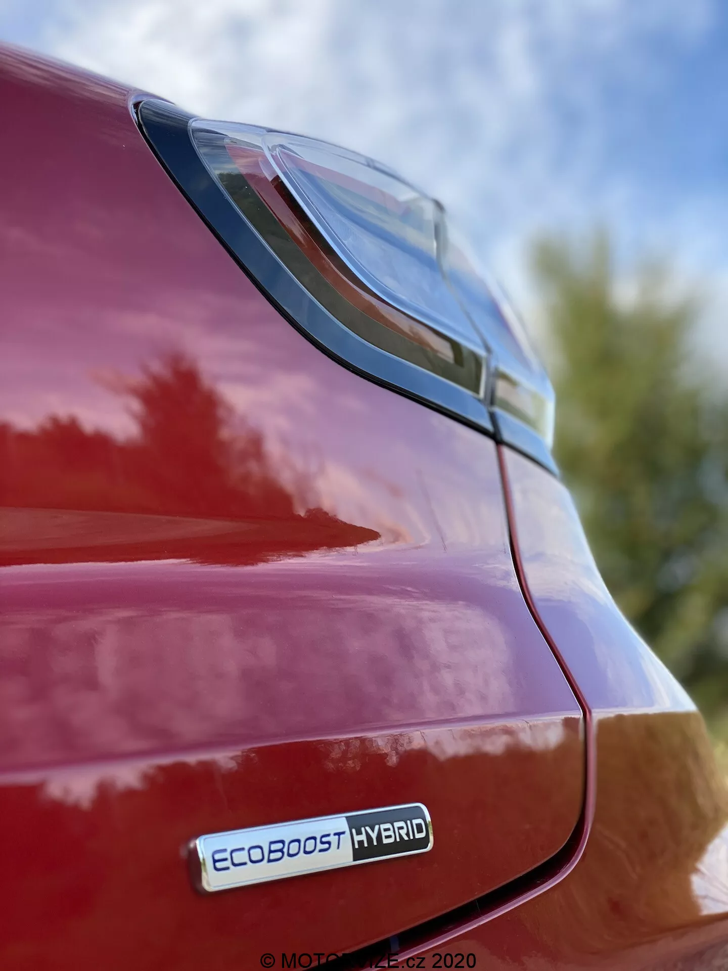 Vista ravvicinata del lato posteriore destro di una Ford Puma rossa 2020, concentrandosi sulla luce posteriore a LED e sull'area del parafango posteriore da una prospettiva a bassa angolazione con il badge EcoBoost Hybrid visibile, riflettendo una finitura lucida e un design elegante contro uno sfondo di cielo e fogliame.