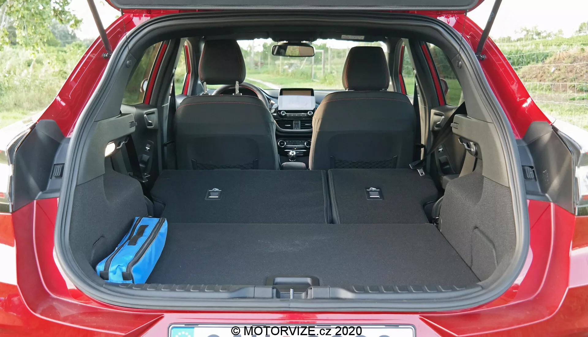 Immagine dell'area di carico della Ford Puma 2020 vista dal retro, con il bagagliaio aperto, i sedili posteriori ribaltati, una borsa blu a destra e un copribagagli retrattile verticale a sinistra, che evidenzia lo spazio di stivaggio adattabile della hatchback