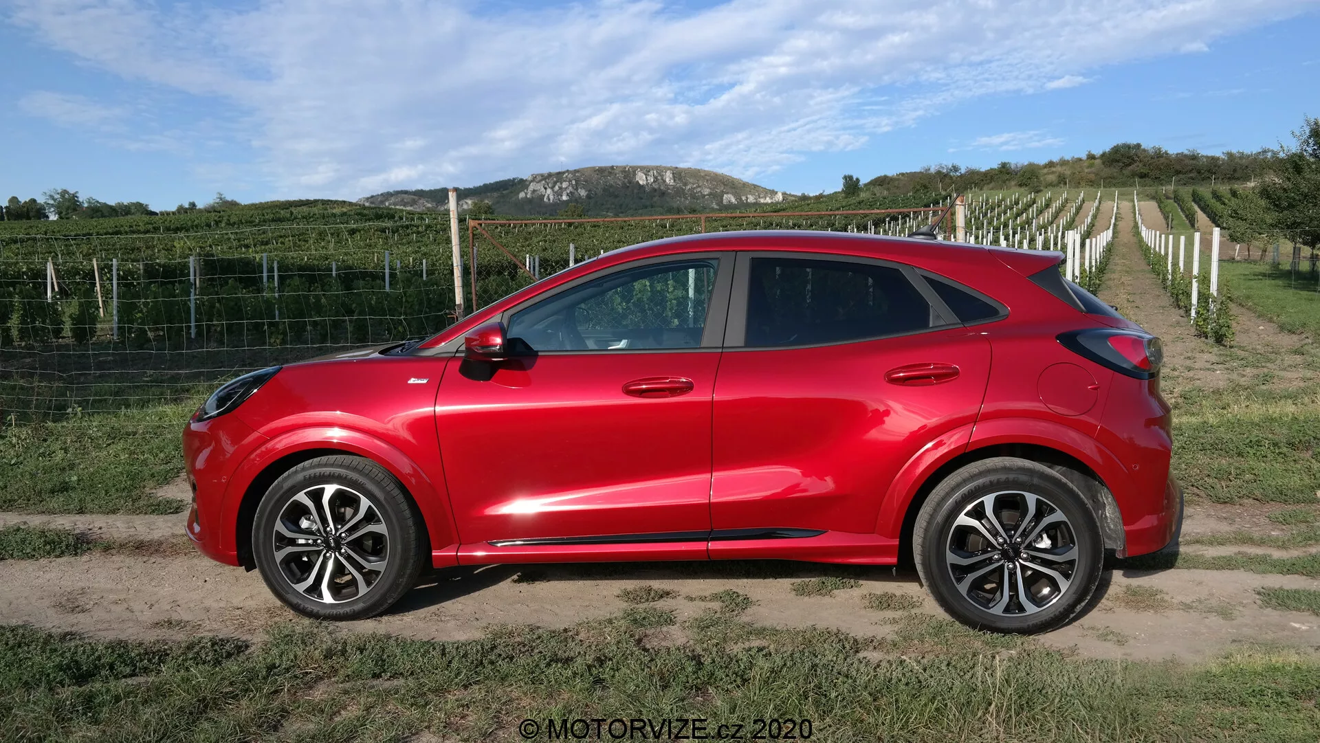 Vista laterale di un SUV crossover compatto Ford Puma 2020 rosso brillante con quattro porte, grandi cerchi in lega, specchietti laterali in tinta carrozzeria con indicatori di direzione integrati, vetri oscurati e maniglie delle portiere in tinta carrozzeria aerodinamiche. Il veicolo, con il suo design aerodinamico moderno e la linea del tetto discendente, è posizionato su un sentiero di terra o ghiaia con un vigneto e cielo sereno sullo sfondo, creando un ambiente rurale.
