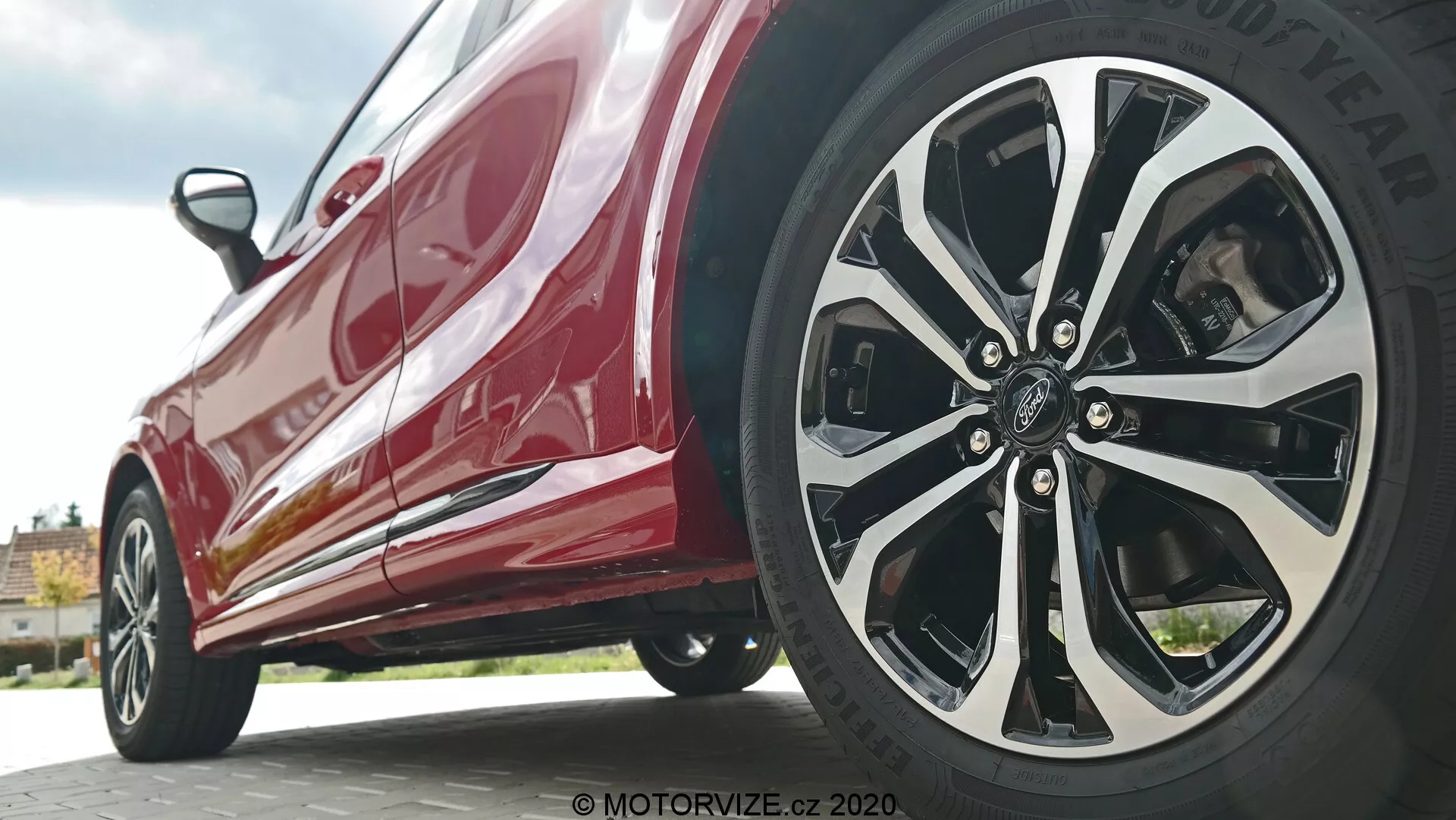 Vista ravvicinata e angolata dal basso della Ford Puma 2020 con messa a fuoco sul profilo laterale vicino alla ruota anteriore. Lo scatto dal livello del suolo mette in evidenza il design a più raggi intricati della ruota del lato del guidatore con finiture miste in argento o cromo e nero, insieme al pneumatico del marchio Goodyear. La carrozzeria rossa lucida riflette la luce, mostrando elementi di design moderni come minigonne laterali e linee aerodinamiche.