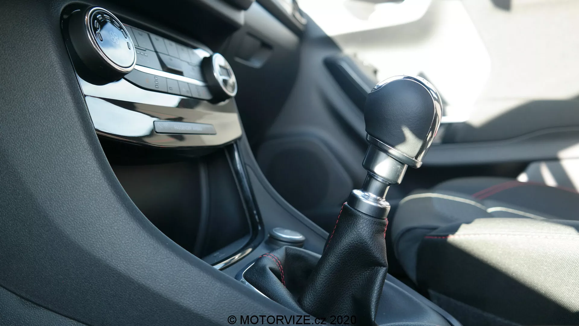Interno di una Ford Puma 2020, concentrandosi sulla console centrale dalla prospettiva del passeggero. L'immagine mette in evidenza una leva del cambio manual a sei velocità in plastica scura con rifiniture in nero lucido attorno alla base. Sullo sfondo è visibile il volante parzialmente sfocato, con pulsanti per il controllo audio e il cruise control, insieme a una bocchetta di ventilazione del cruscotto. La scena è illuminata dalla luce naturale del giorno.