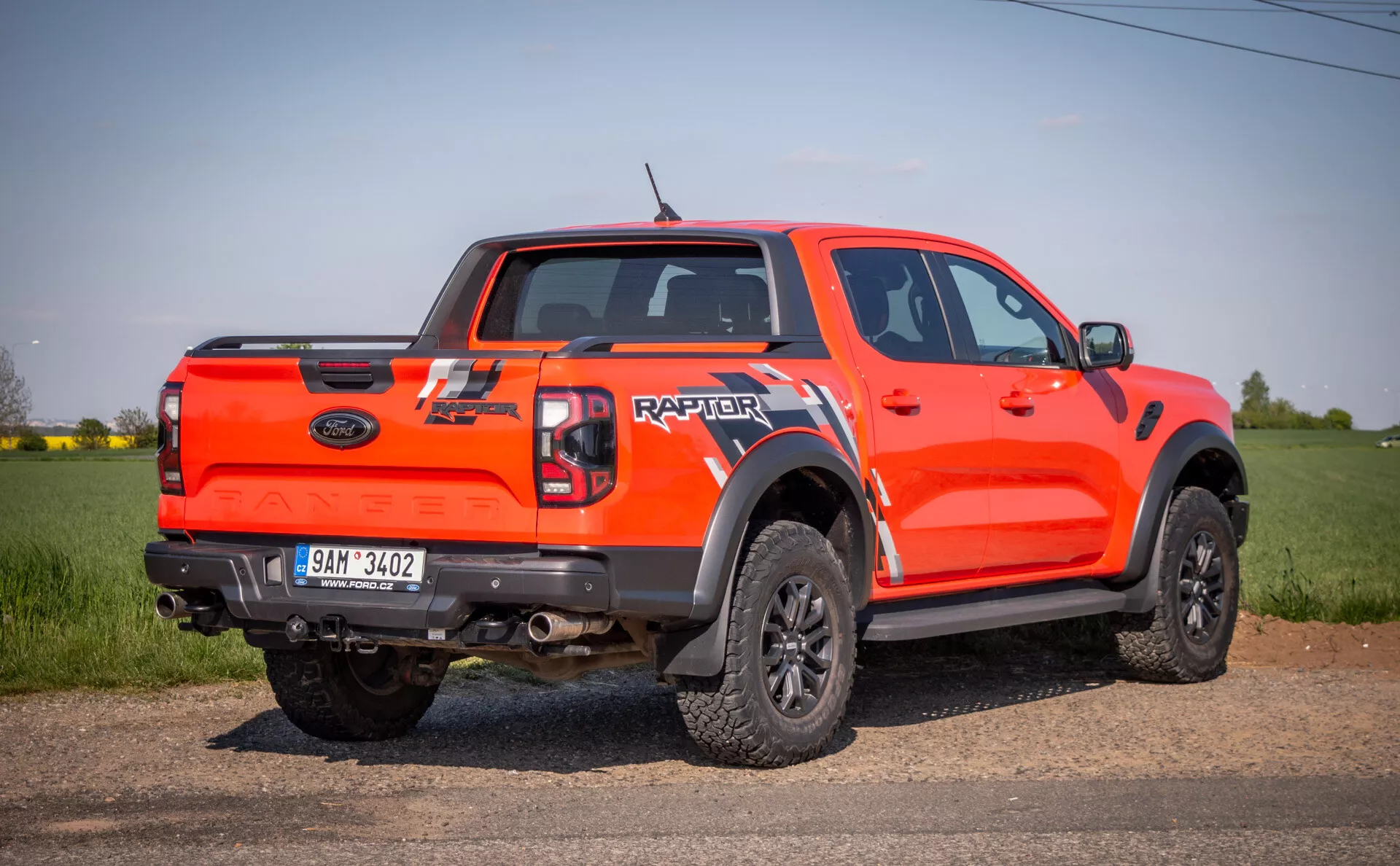 Camioncino Ford Raptor 2022 arancione brillante in prospettiva laterale-posteriore, all'aperto su un campo, che mette in mostra le sue capacità fuoristrada con elevata altezza da terra, cabina a quattro porte, ampie estensioni degli archi ruota neri, ruote nere a più razze con pneumatici da off-road, e il marchio 'RAPTOR' sul portellone posteriore.