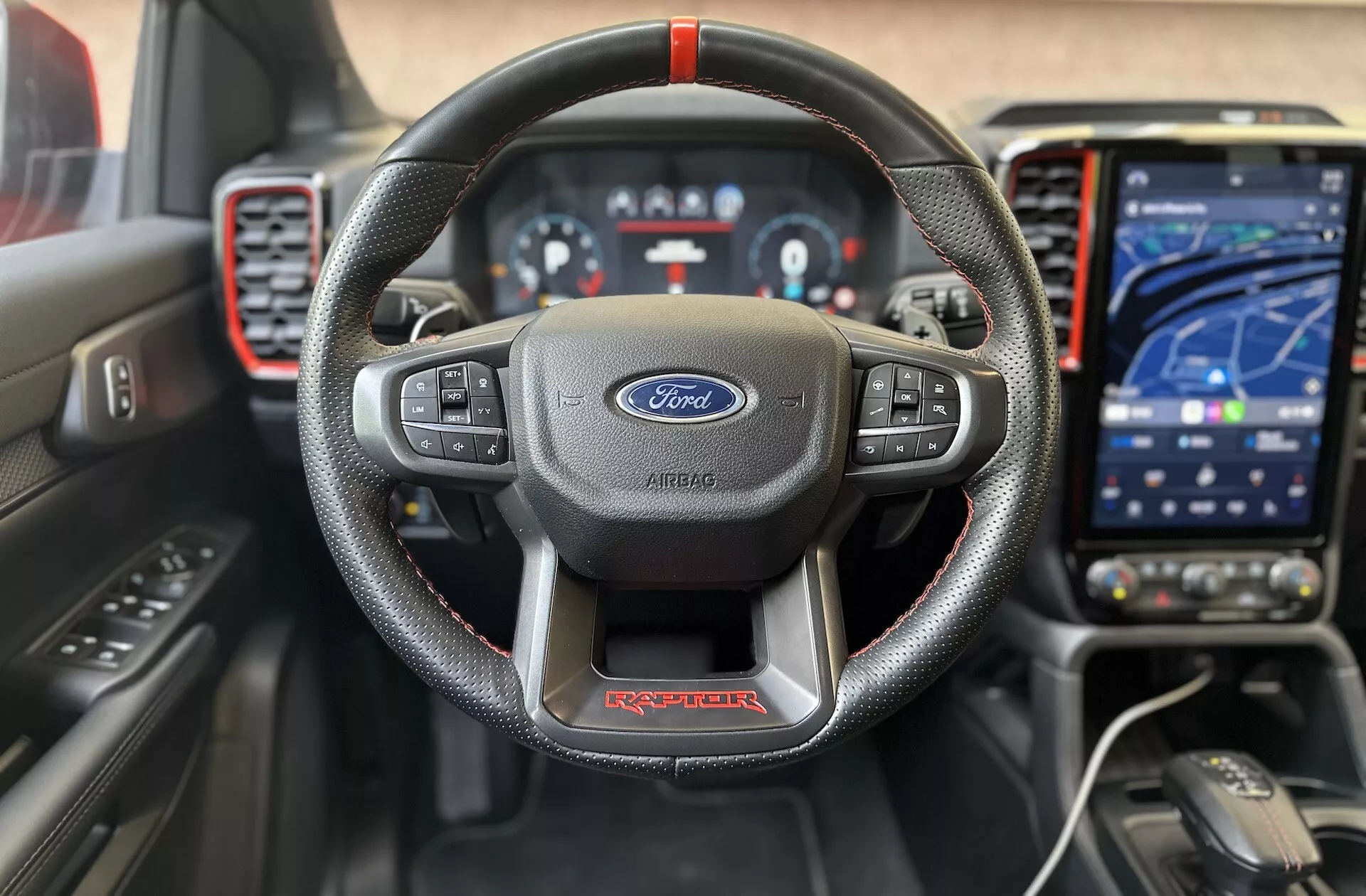 Vista frontale interna del Ford Ranger Raptor 2022 che evidenzia il volante a tre razze avvolto in pelle con cuciture rosse e logo Ford, quadro strumenti digitale, moderno sistema di infotainment touch in modalità navigazione, cruscotto sportivo con accenti rossi e aiuti avanzati per il conducente per un'esperienza di guida di alta qualità e focalizzata sulla tecnologia