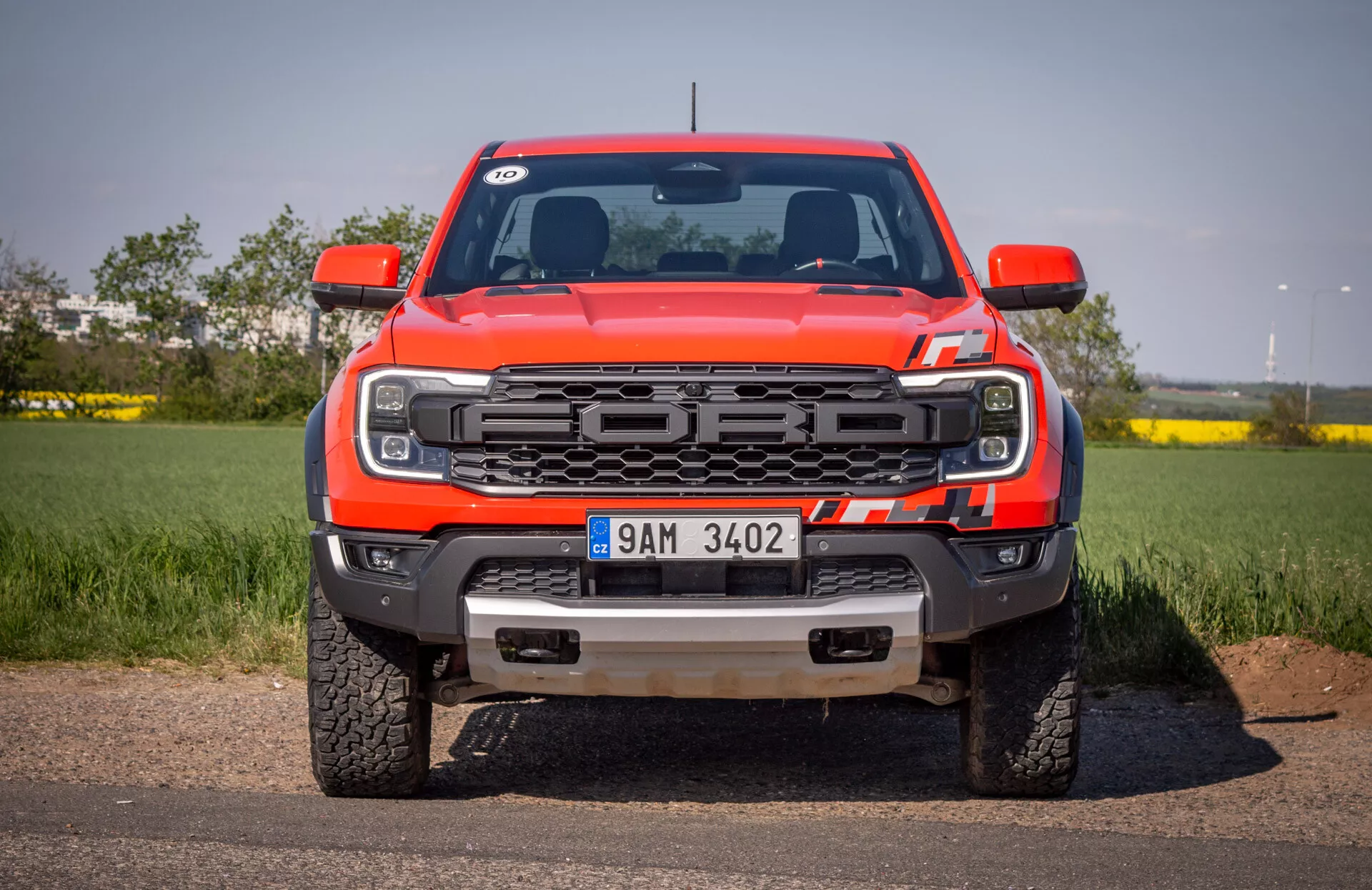 Vista frontale del pickup rosso Ford Ranger 2022 sull'asfalto con sfondo di cielo sereno, caratterizzata da griglia, fari LED, paraurti anteriore con ganci di traino e piastra di protezione, e logo Ford