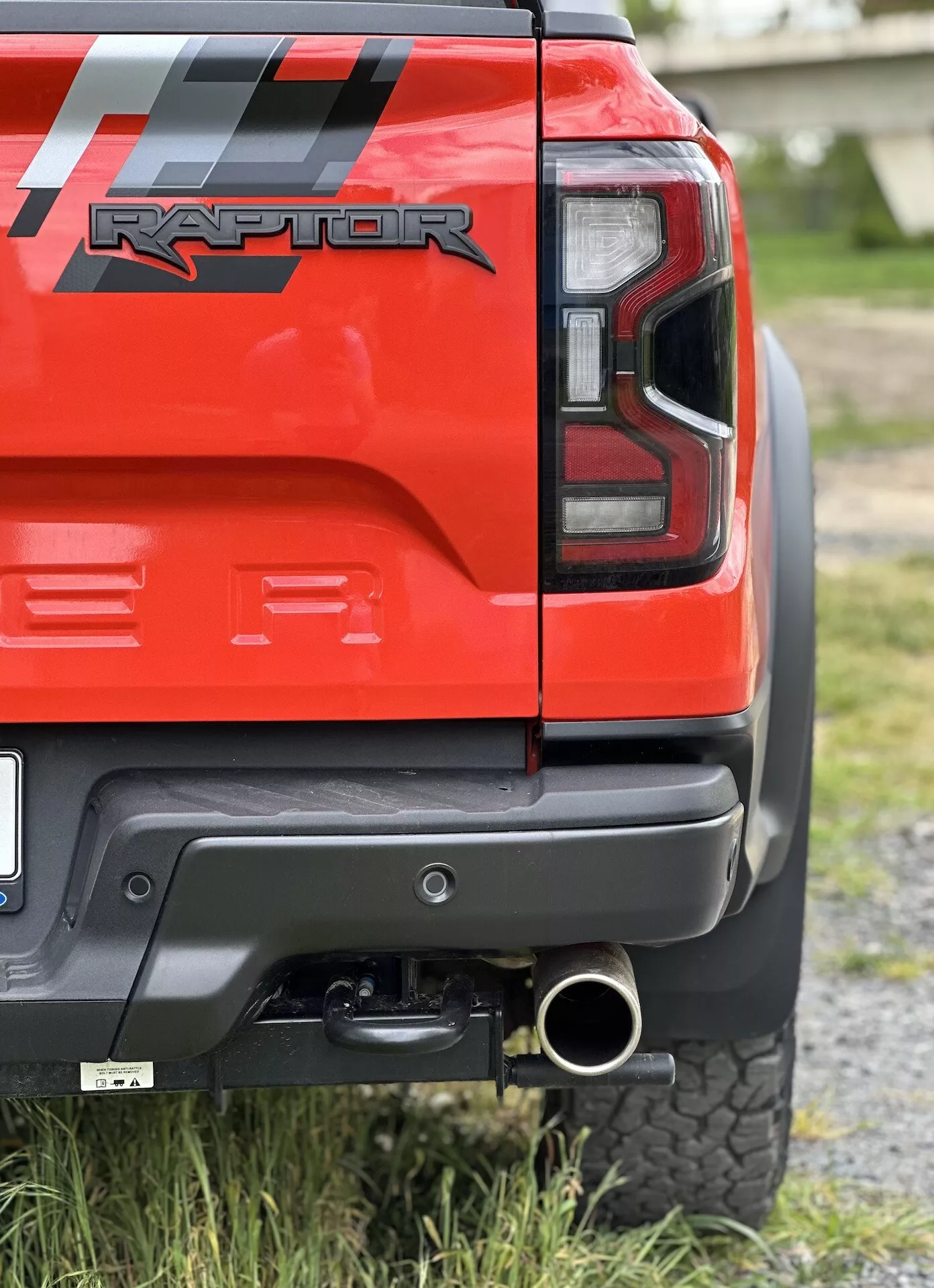 Primo piano dell'angolo posteriore di un Ford Ranger Raptor rosso del 2022 che mostra la luce posteriore a LED, il paraurti posteriore nero con pannello grigio, il gancio di traino, la punta dello scarico lucidata e il pneumatico all-terrain, sottolineando le sue capacità off-road e i dettagli di design.