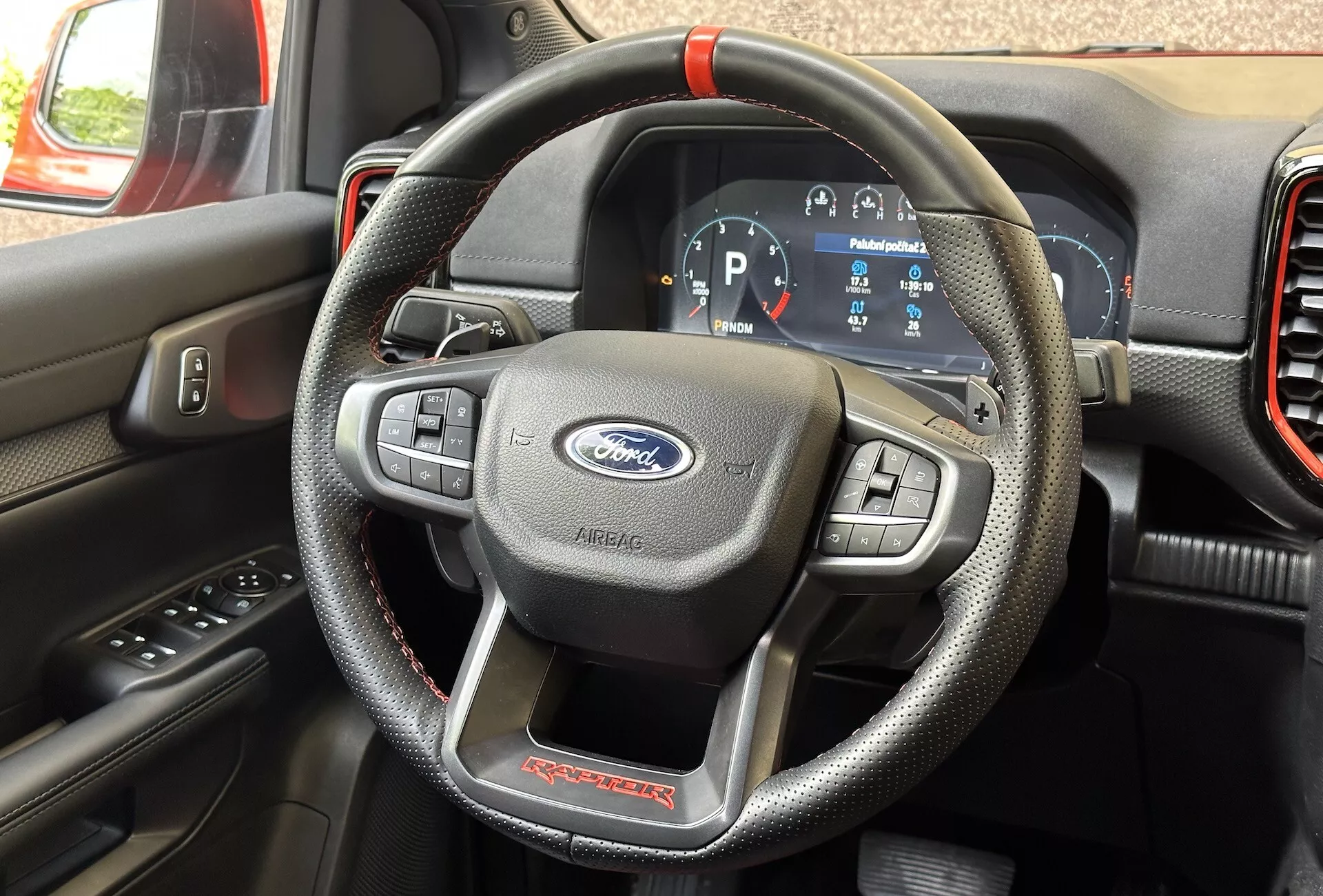 Vista dettagliata dell'interno di un Ford Ranger Raptor 2022 focalizzata sul volante in materiale simil-pelle con cuciture rosse e comandi multifunzione, e il cruscotto digitale che mostra il veicolo in modalità Parcheggio.