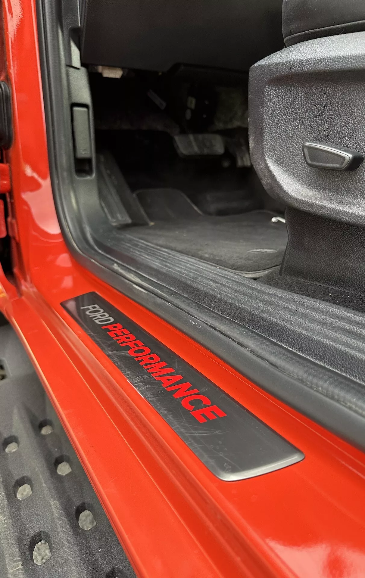 Primo piano della soglia della porta del Ford Ranger 2022 con distintivo 'FORD PERFORMANCE', esterno rosso lucido, tappeto interno nero, sedile in tessuto grigio dal design sportivo e tappetino in gomma nera resistente