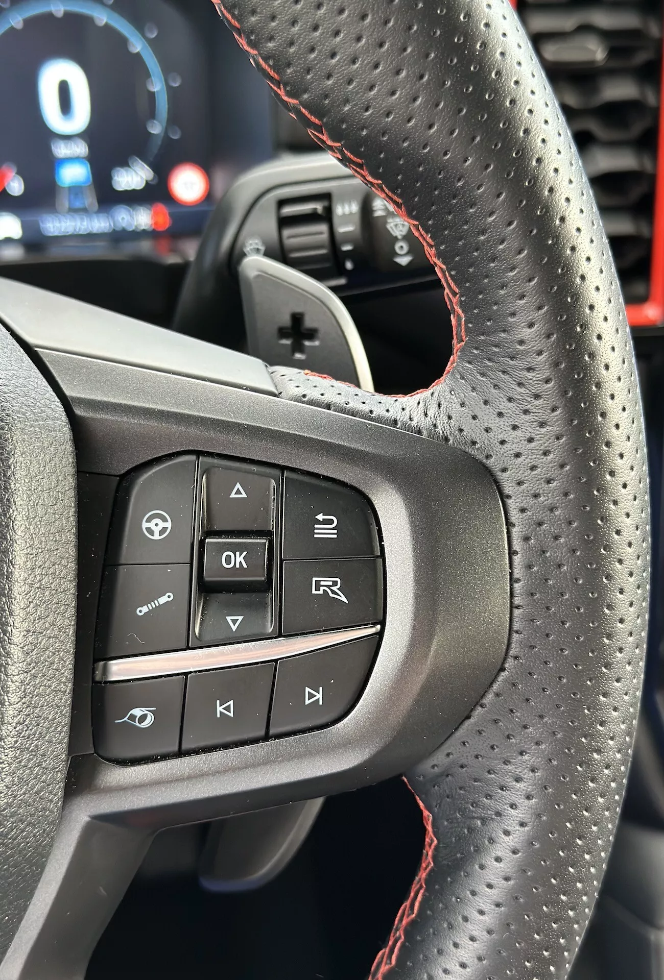 Primo piano del volante del Ford Ranger 2022 che evidenzia i pulsanti e le caratteristiche sul lato sinistro, inclusa la pelle forata con cuciture rosse, dalla prospettiva del conducente in orientamento verticale