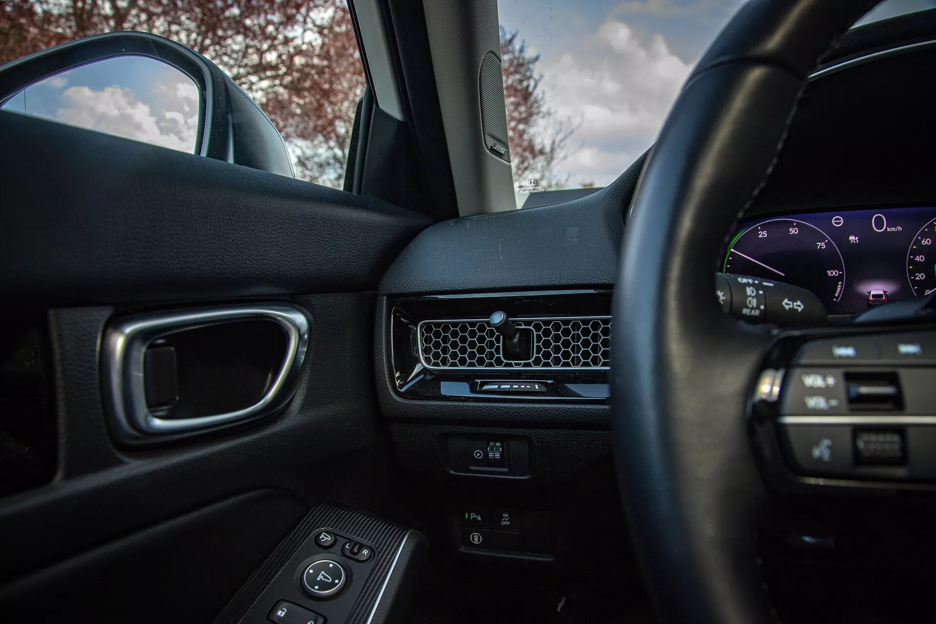 Vista interna del Honda Civic 2022 dal sedile del conducente che mostra volante con comandi multimediali, cruscotto minimalista con bocchetta a nido d'ape, quadro strumenti digitale, porta del passeggero con comandi per i finestrini elettrici e sedile del passeggero anteriore di colore scuro con illuminazione naturale