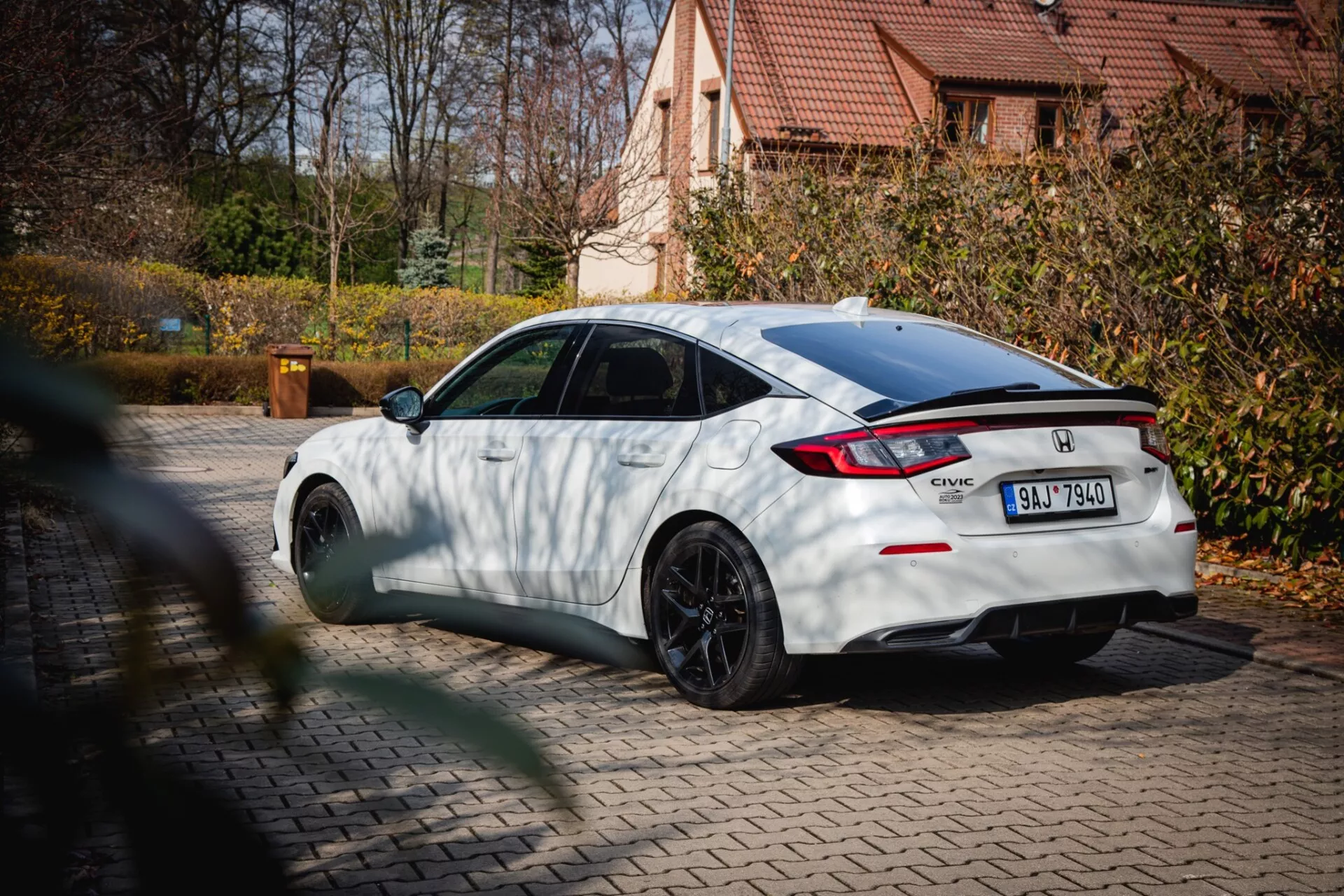 Honda Civic bianca del 2022 con ruote multirazze nere e spoiler posteriore, parcheggiata su una pavimentazione con fogliame autunnale e una casa residenziale sullo sfondo