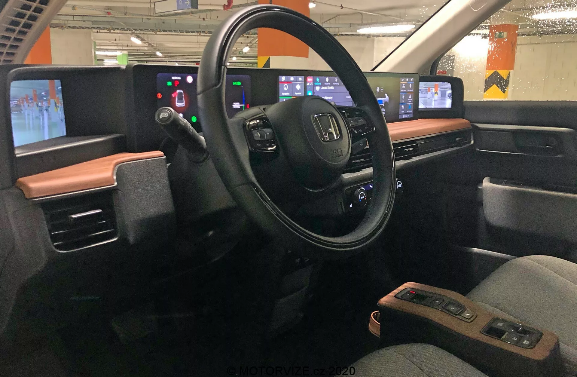 Vista interna della Honda e 2020, focalizzata sull'area del conducente vista dal centro del veicolo. Il cruscotto presenta un design moderno con uno schema di colori a due tonalità. Il volante, con il logo Honda e vari pulsanti di controllo, è al centro, circondato da un pannello digitale invece di indicatori analogici. A destra, la consolle centrale digitale include pannelli touchscreen per audio, navigazione, controllo del clima e altre impostazioni. Il design enfatizza l'integrazione tecnologica, minimizzando i pulsanti fisici a favore delle interfacce touchscreen. L'auto sembra essere ferma in un parcheggio, con luci fluorescenti e pilastri in cemento visibili attraverso i finestrini.