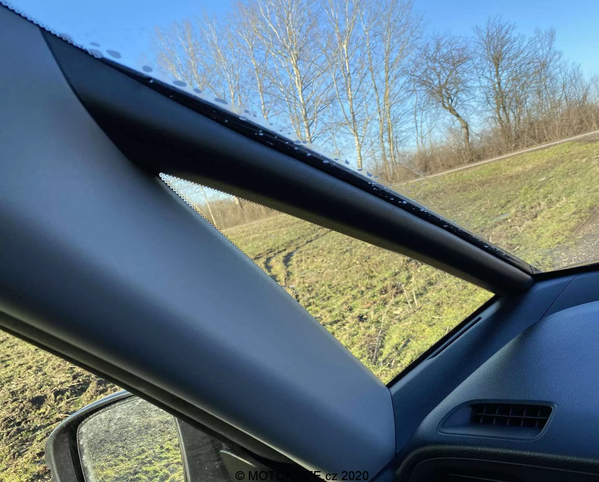 Vista interna di una Honda Jazz Crosstar 2020 guardando verso il finestrino lato guidatore, con un focus sul montante A. Il montante A è ricoperto di un materiale non metallico e testurizzato e ostruisce parzialmente la visuale. Il finestrino laterale mostra gocce d'acqua, indicanti pioggia recente o condensa, con un'area erbosa e alberi visibili all'esterno sotto un cielo azzurro e limpido.