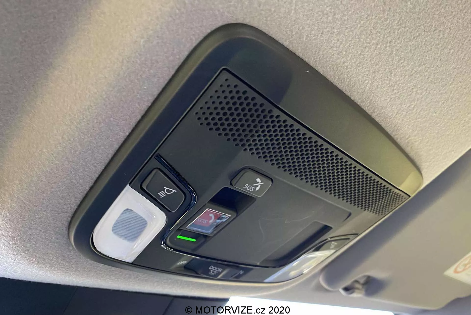 Vista ravvicinata della console sopraelevata della Honda Jazz 2020 (Jazz Crosstar). L'immagine rivela una serie di pulsanti funzionali, tra cui controlli delle luci interne, un possibile pulsante di sblocco della porta, un'area ventilata con microfono per chiamate in vivavoce, un home link con pulsanti programmabili per l'apertura del garage e un possibile vano portaocchiali o fermaglio di stoccaggio. La console è catturata alla luce del giorno con una messa a fuoco nitida sui pulsanti texturizzati, sottolineando la tecnologia avanzata e il design degli interni del veicolo.