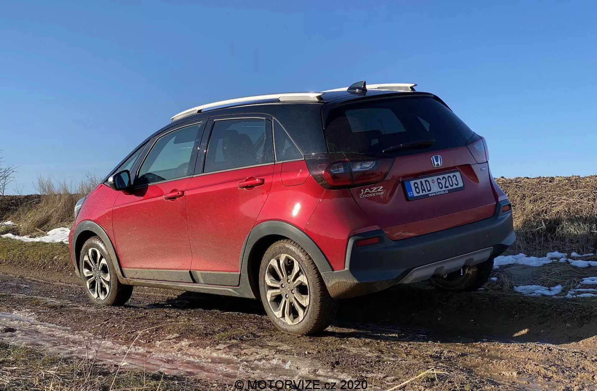 Un SUV Honda Jazz Crosstar rosso del 2020 visto da una prospettiva posteriore di tre quarti, che mostra dettagli come emblemi di marca e modello, luci posteriori a LED, paraurti posteriore con graffi e sensori, cerchi in lega, pneumatici per tutte le stagioni, barre sul tetto, antenna a pinna di squalo, finestrini oscurati con linee di sbrinamento e targa centrata. L'apertura del bagagliaio di tipo hatchback e le macchie di neve sul terreno con un campo asciutto sullo sfondo indicano un'atmosfera di fine autunno, inverno o inizio primavera sotto un cielo limpido a mezzogiorno o nel primo pomeriggio.