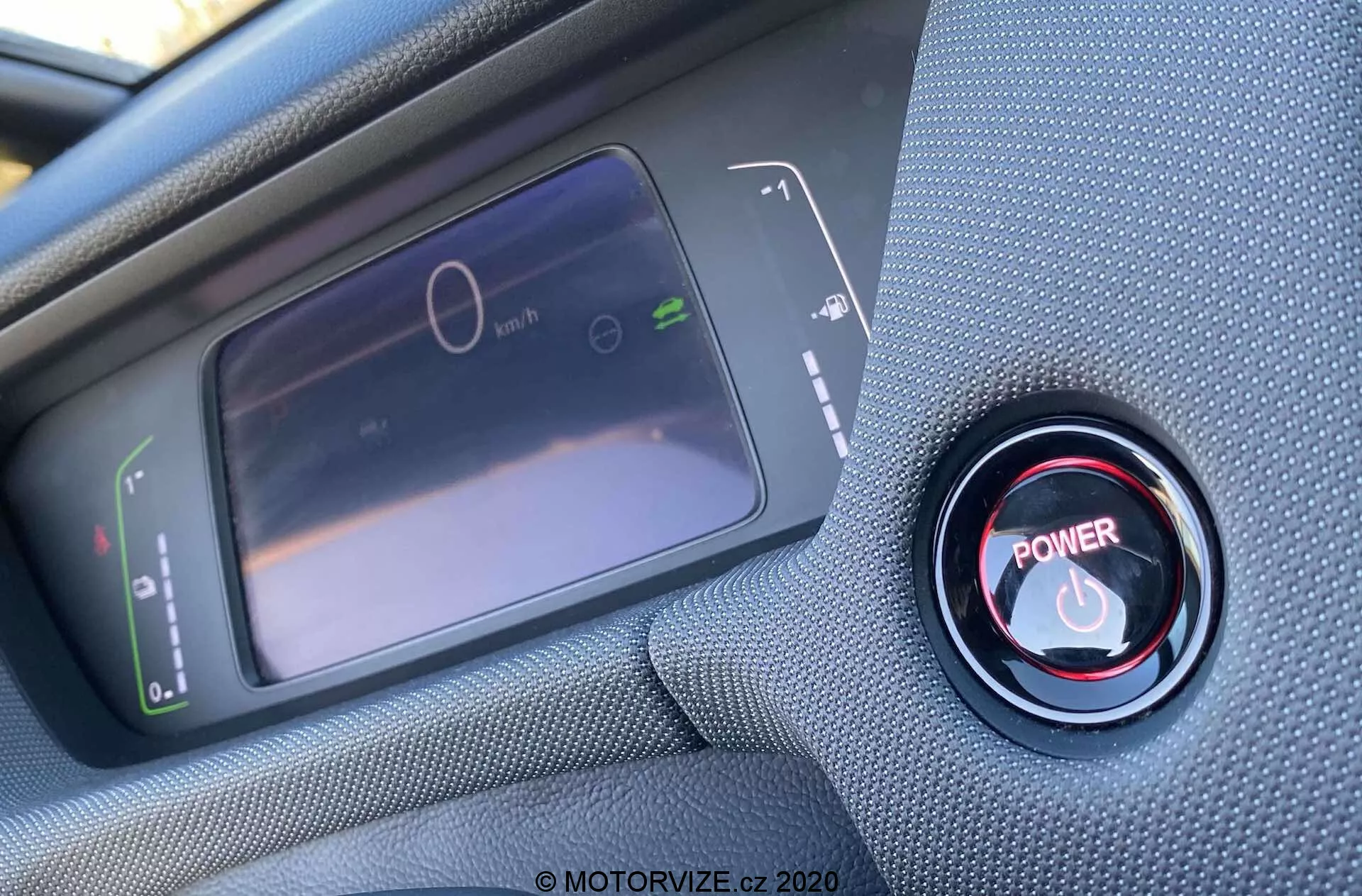 Vista interna ravvicinata dal punto di vista del conducente di una Honda Jazz Crosstar 2020, con particolare attenzione al display del tachimetro digitale e al pulsante rosso di avviamento/arresto del motore. Il display LCD a sinistra mostra un tachimetro fermo e un grafico del consumo energetico, mentre a destra, il pulsante rosso di avviamento/arresto è circondato da un anello metallico. La foto è scattata da un angolo obliquo dall'alto, con una messa a fuoco nitida sul pulsante e una messa a fuoco morbida sul display.