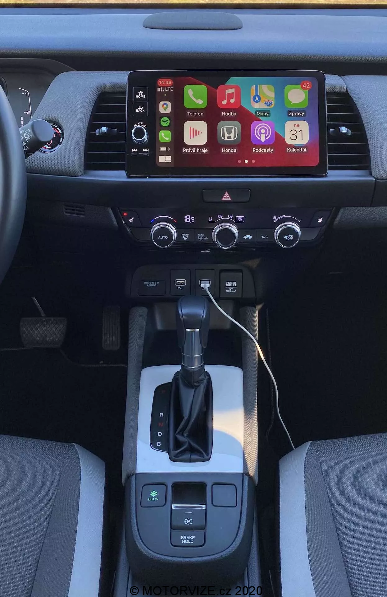 Vista interna della console centrale della Honda Jazz Crosstar 2020 da un'angolazione leggermente elevata e arretrata. Si concentra sull'area del cambio, selettore del cambio automatico e touchscreen infotainment con diverse icone di app, suggerendo opzioni di connettività avanzate come Apple CarPlay e Android Auto. Sotto lo schermo ci sono le prese d'aria e un pannello di controllo del clima a doppia zona con pulsanti e manopole fisici. La leva del cambio è affiancata dal selettore della modalità di guida e dai pulsanti del freno di stazionamento elettronico, con uno spazio di archiviazione aperto a destra che indica le porte di ricarica. I sedili anteriori bicolore evidenziano il design interno e il comfort.
