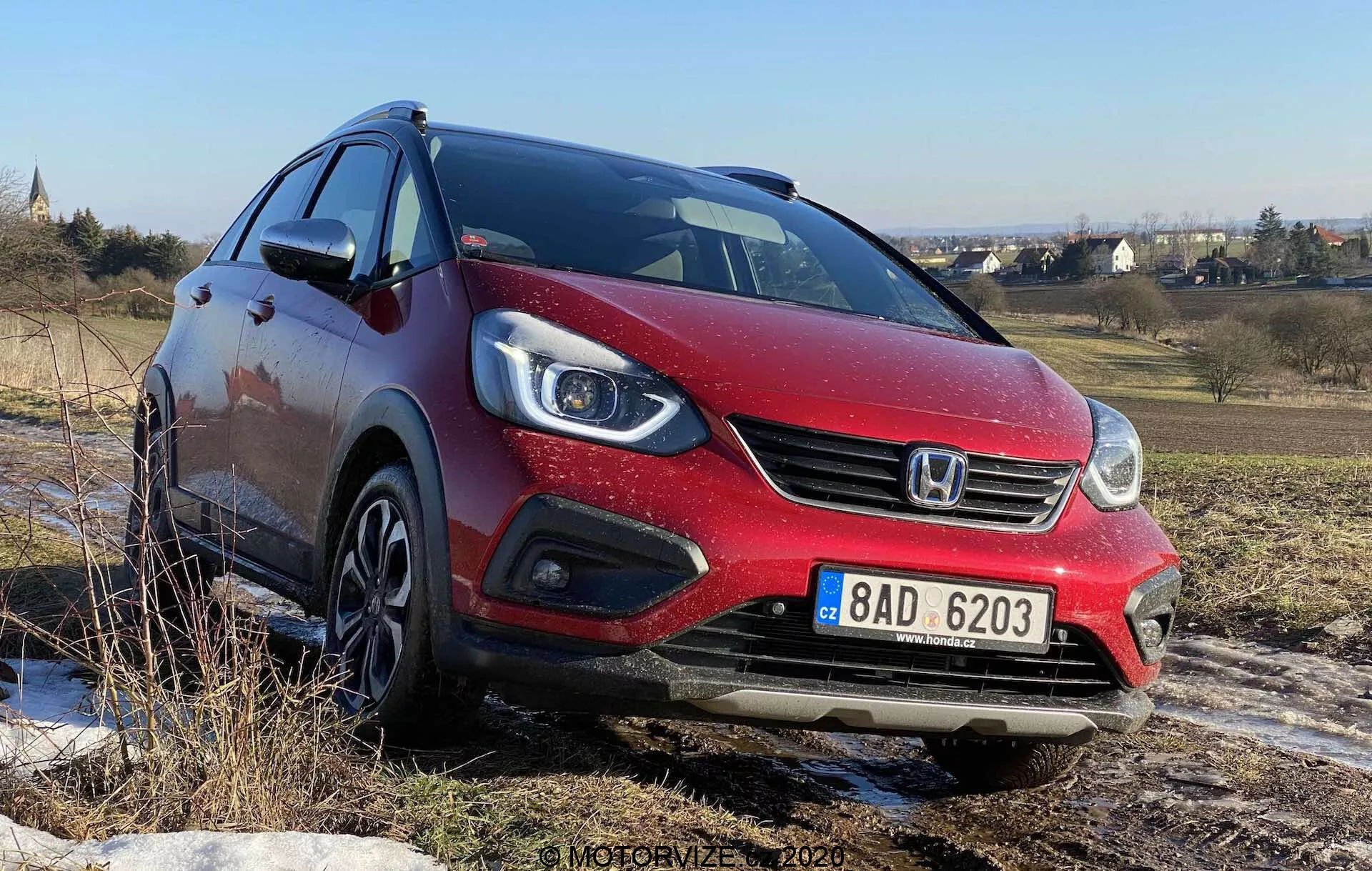 Vista frontale tre quarti di una Honda Jazz Crosstar 2020 rossa, ripresa da un angolo basso, che mostra la sua fascia anteriore, fari alogeni a proiettore con luci diurne a LED integrate, griglia anteriore con logo del marchio e cerchi in lega. L'auto è su un terreno accidentato sotto un cielo azzurro, suggerendo la sua capacità di gestire superfici non asfaltate.