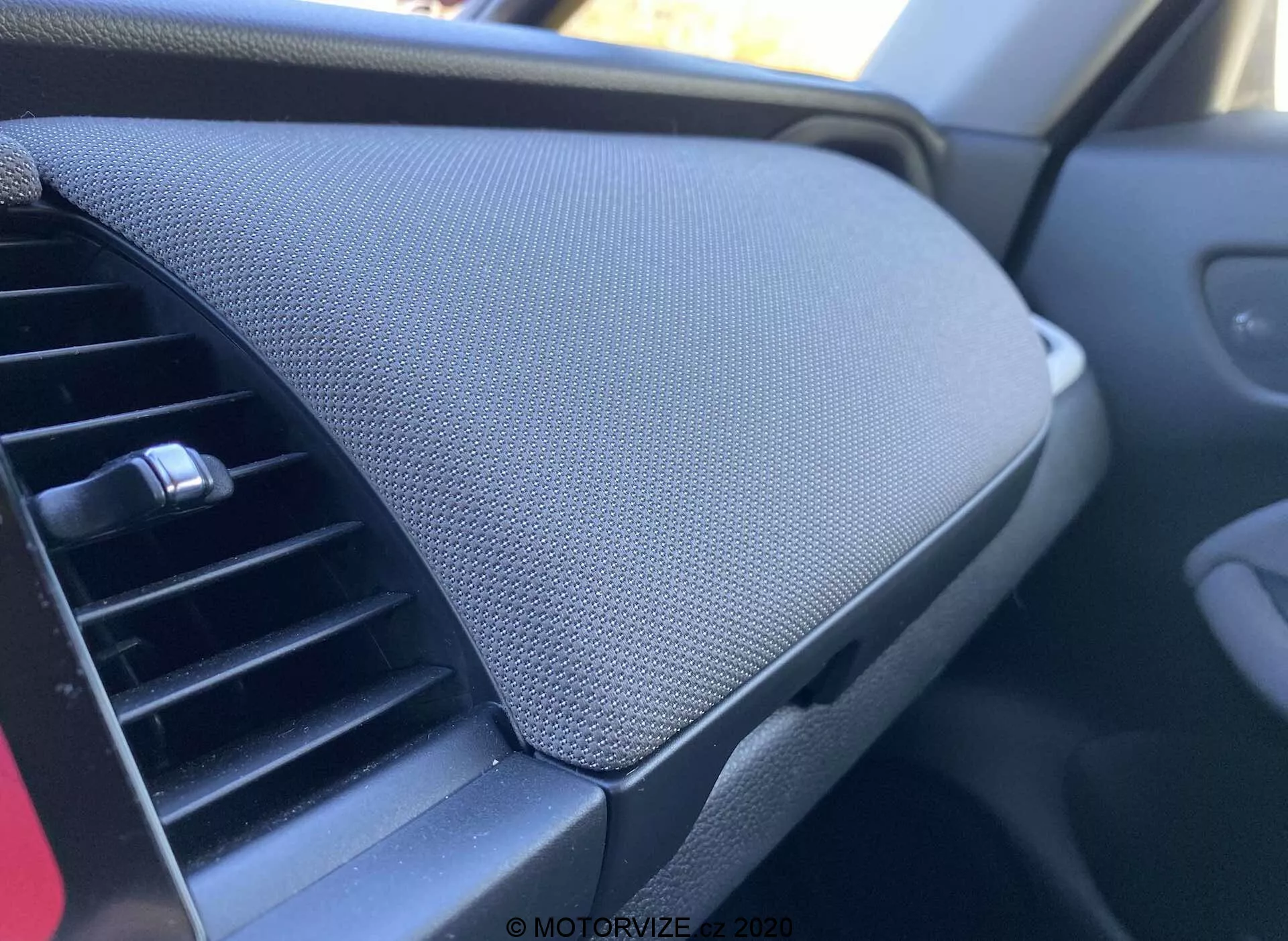 Vista ravvicinata dell'interno di una Honda Jazz 2020 (Jazz Crosstar), concentrata sul cruscotto. Presa dalla prospettiva del passeggero, l'immagine mostra la bocchetta dell'aria condizionata a sinistra, con un design orizzontale regolabile. Dominante a destra dell'immagine è il vano portaoggetti del cruscotto con coperchio chiuso, caratterizzato da una superficie testurizzata, forse in rete o tessuto. Il vano sembra integrato senza una maniglia visibile, suggerendo un meccanismo di apertura a pressione. Il schema di colori complessivo è scuro, implicando un'estetica neutra mirata a ridurre le distrazioni.