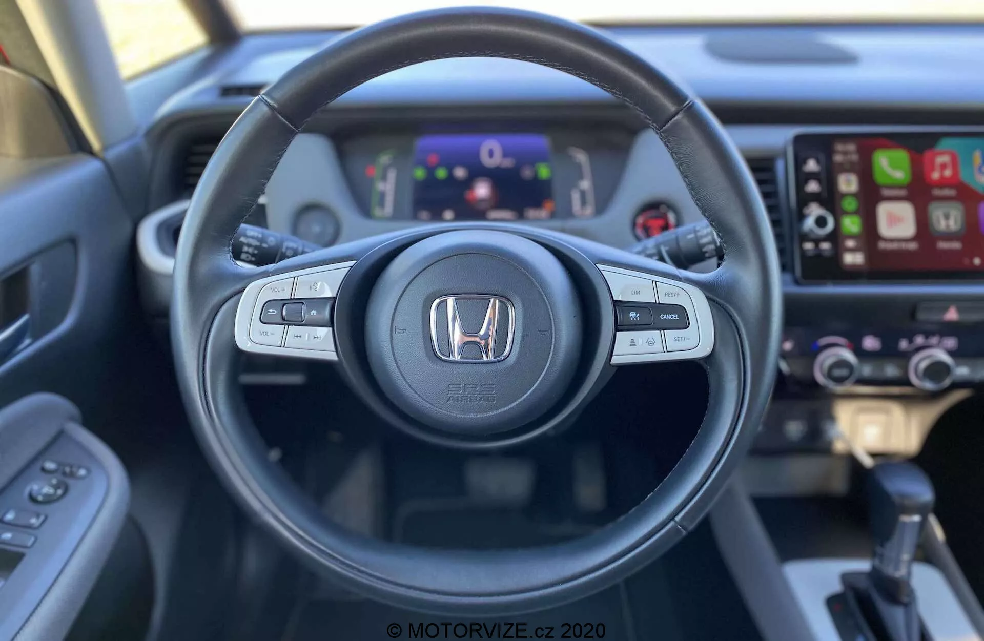 Vista dell'interno della Honda Jazz Crosstar 2020 dal sedile del conducente, con un volante dotato di pulsanti per controllo audio, controllo vocale e controllo di crociera. Il logo Honda è ben visibile al centro. Dietro il volante, un quadro strumenti digitale mostra vari indicatori e informazioni. A destra, è visibile un sistema di infotainment touchscreen compatibile con Apple CarPlay, insieme a controlli del clima leggermente sfocati. L'immagine, scattata da una prospettiva in prima persona, mette in evidenza il design ergonomico e l'accessibilità della tecnologia del veicolo.