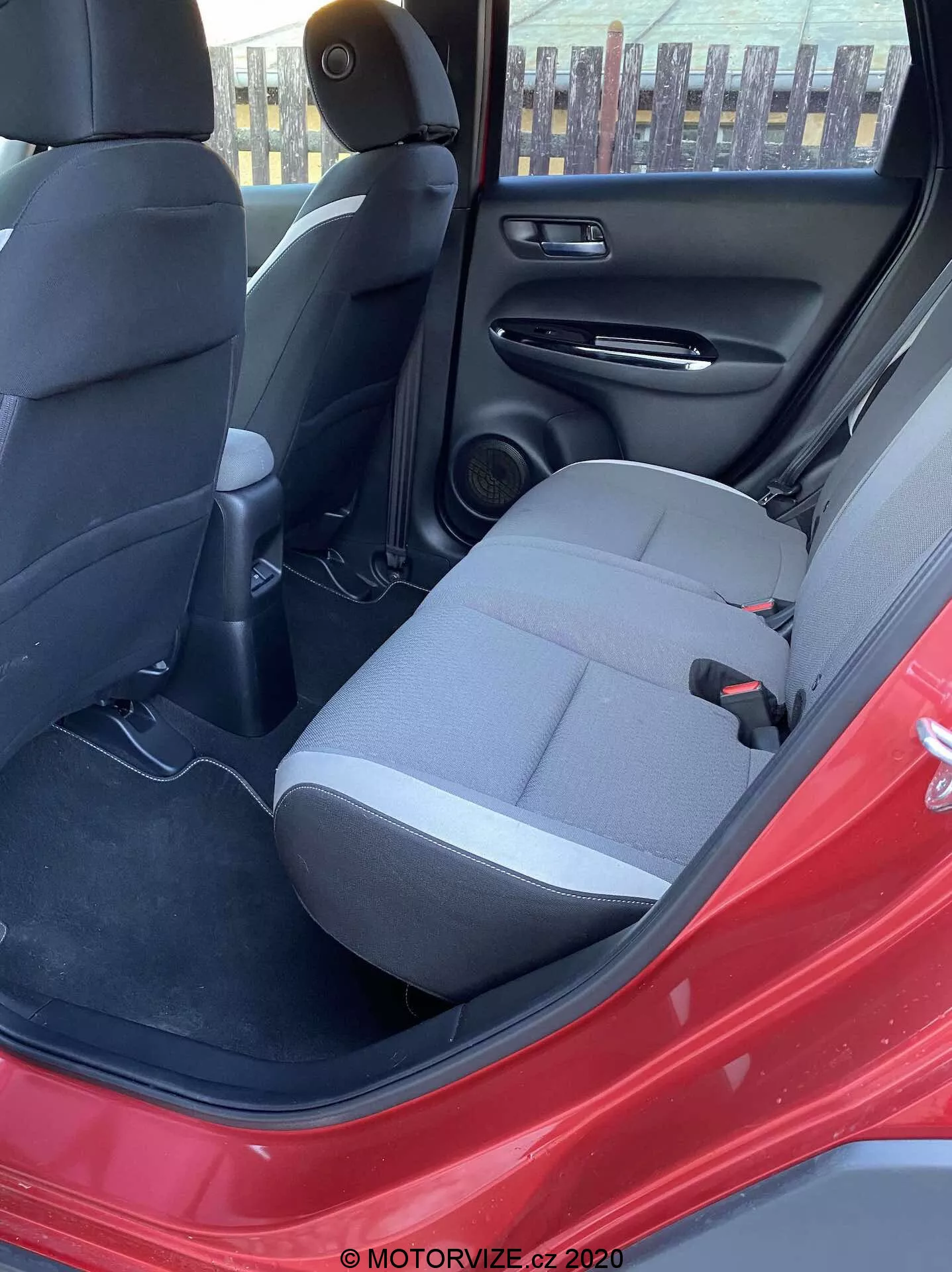 Vista dell'interno della Honda Jazz Crosstar 2020 dalla porta posteriore lato guida aperta, evidenziando il sedile passeggero posteriore in tessuto scuro, cintura di sicurezza, console centrale posteriore con bocchette d'aria, altoparlante nella portiera posteriore, pannello interno nero con tasca portaoggetti, tappetino sul pavimento e il vivace colore rosso dell'esterno dell'auto. La luce naturale del giorno illumina la scena.