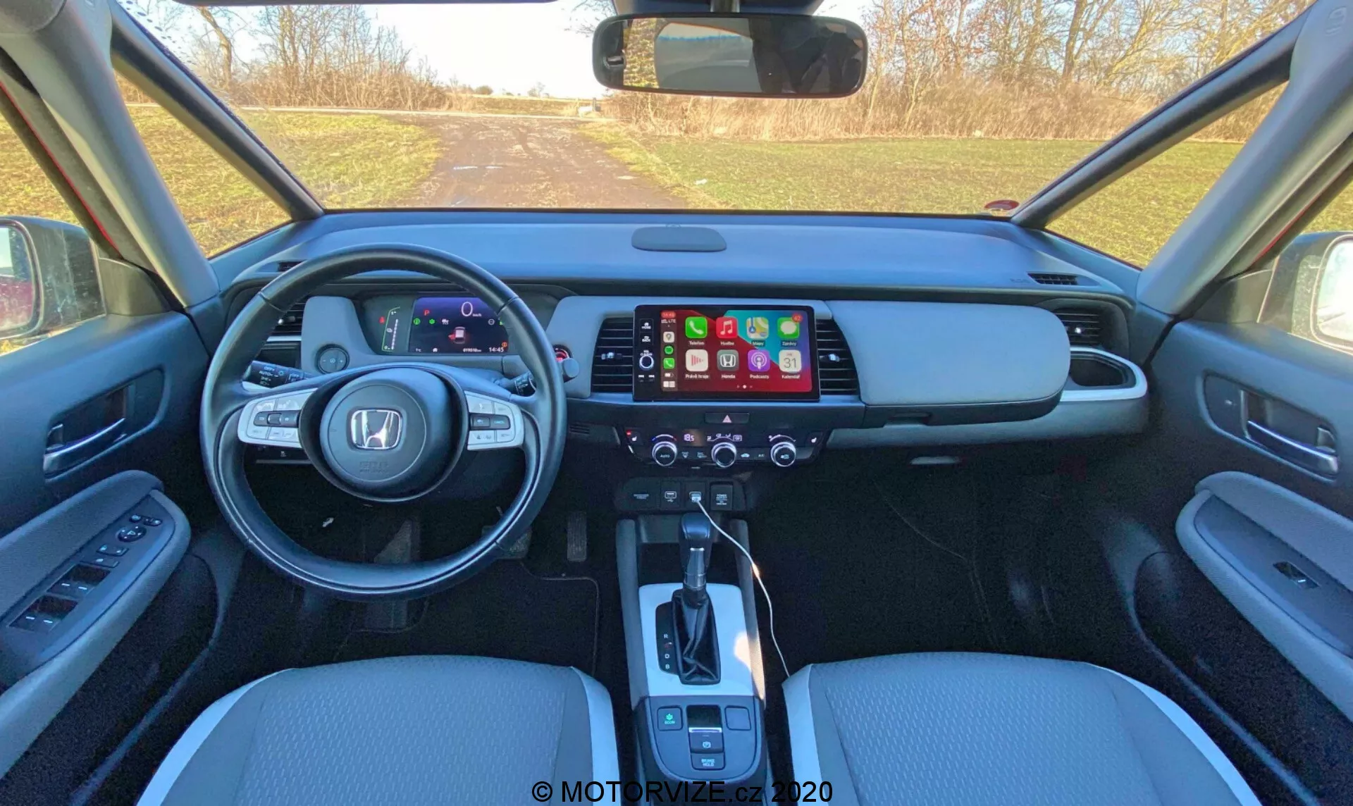 Vista interna della Honda Jazz 2020 (Jazz Crosstar) osservata dall'area dei sedili posteriori, che mostra il cruscotto anteriore con quadro strumenti digitale, un ampio schermo touchscreen dell'infotainment, pannello di controllo del clima, leva del cambio automatico, sedili in pelle e comandi del pannello porta.