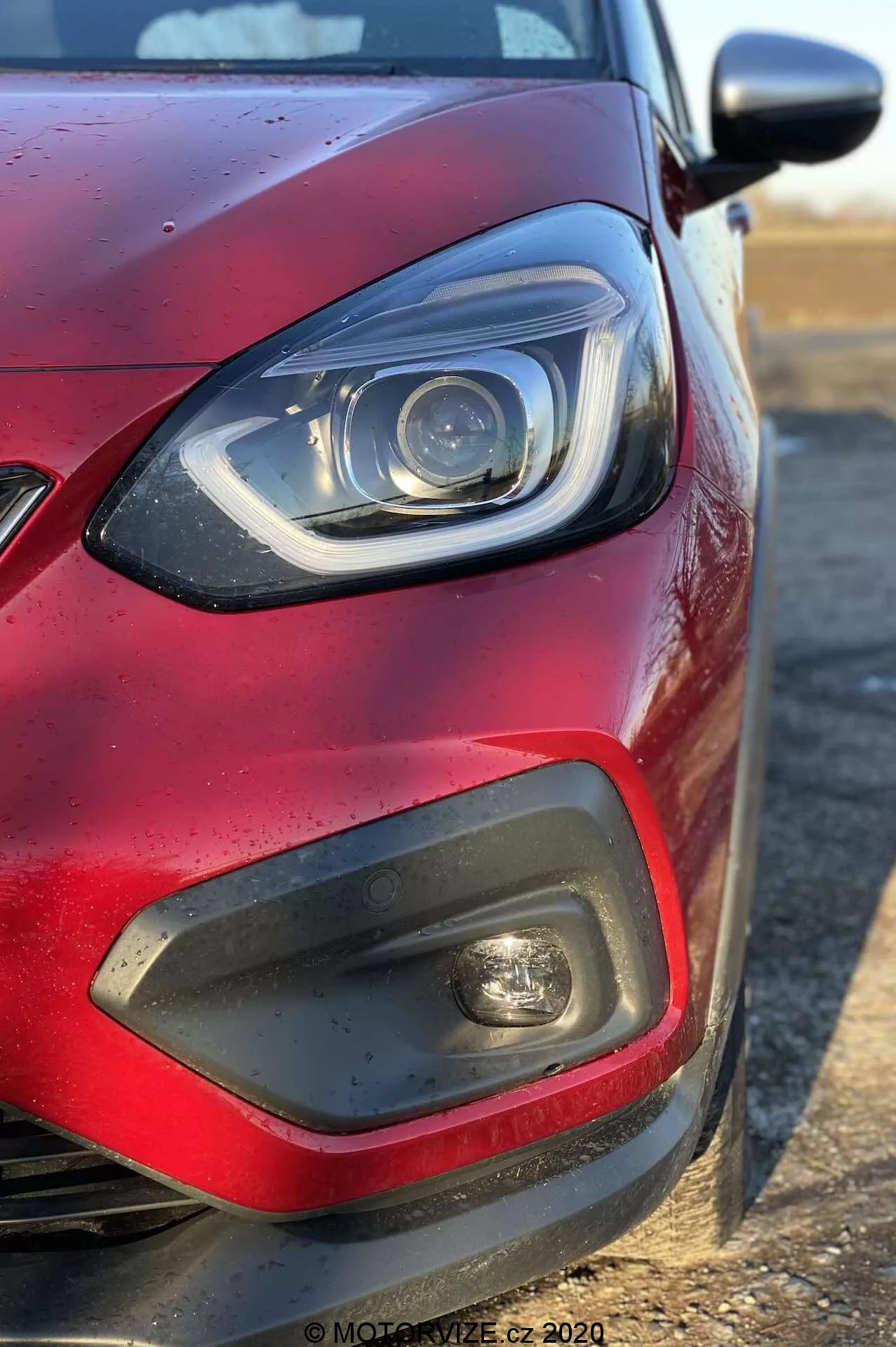 Inquadratura ravvicinata del quadrante anteriore sinistro di una Honda Jazz 2020 (Jazz-Crosstar) rossa da un'angolazione laterale, concentrandosi sul faro con elementi LED o xenon e una barra DRL curva, un inserto in plastica nera con fendinebbia, una parte della griglia, vernice rossa metallizzata con gocce d'acqua e uno specchietto laterale dello stesso colore della carrozzeria che si estende da destra.