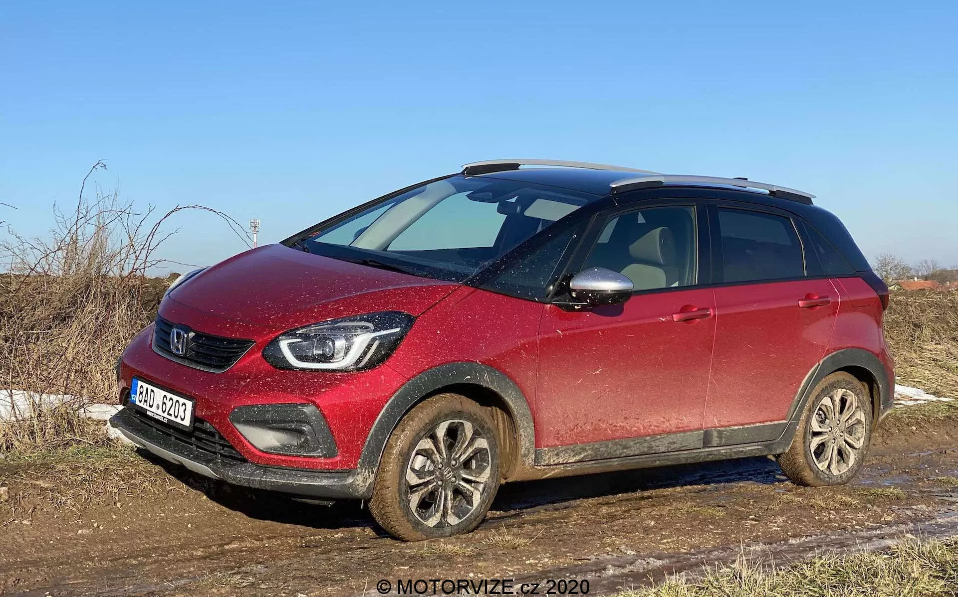Vista frontale a tre quarti di un SUV crossover compatto Honda Jazz Crosstar 2020 rosso, catturato alla luce del giorno dal lato passeggero, che mostra la griglia frontale, i fari, i cerchi in lega ricoperti di fango e il profilo laterale fino alla portiera posteriore passeggero. L'auto appare leggermente sporca, indicando un uso recente su strade non asfaltate o bagnate. Le caratteristiche visibili includono la griglia prominente con il logo del marchio, fari LED moderni, fari fendinebbia, cerchi in lega, specchietti laterali con indicatori di direzione integrati, barre del tetto e maggiore altezza da terra, suggerendo la versatilità e la capacità del veicolo per un uso leggero fuoristrada. L'SUV è parcheggiato in un ambiente rurale con erba secca e cieli sereni, evidenziando la sua idoneità per le strade di campagna e il fuoristrada leggero.