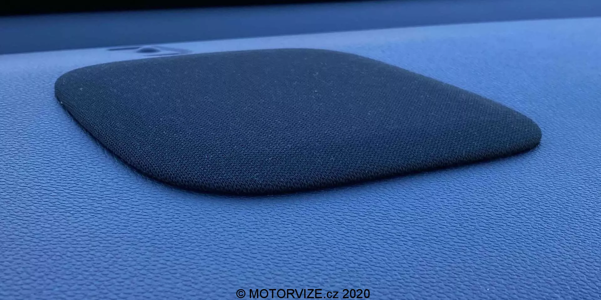 Fotografia ravvicinata di una griglia del diffusore dell'auto incastonata in un cruscotto o pannello porta di colore blu scuro o nero, probabilmente della Honda Jazz 2020. La griglia del diffusore è realizzata con un materiale testurizzato simile al tessuto e ha una forma di trapezio troncato o quadrilatero irregolare con un lato leggermente convesso. Il componente è leggermente rialzato, proiettando un'ombra che enfatizza il suo profilo e la sua texture. La foto è scattata con un'angolazione laterale utilizzando luce naturale diffusa, mettendo in risalto la texture granulosa della superficie circostante e contiene una filigrana che recita '© MOTORVIZE.cz 2020.'