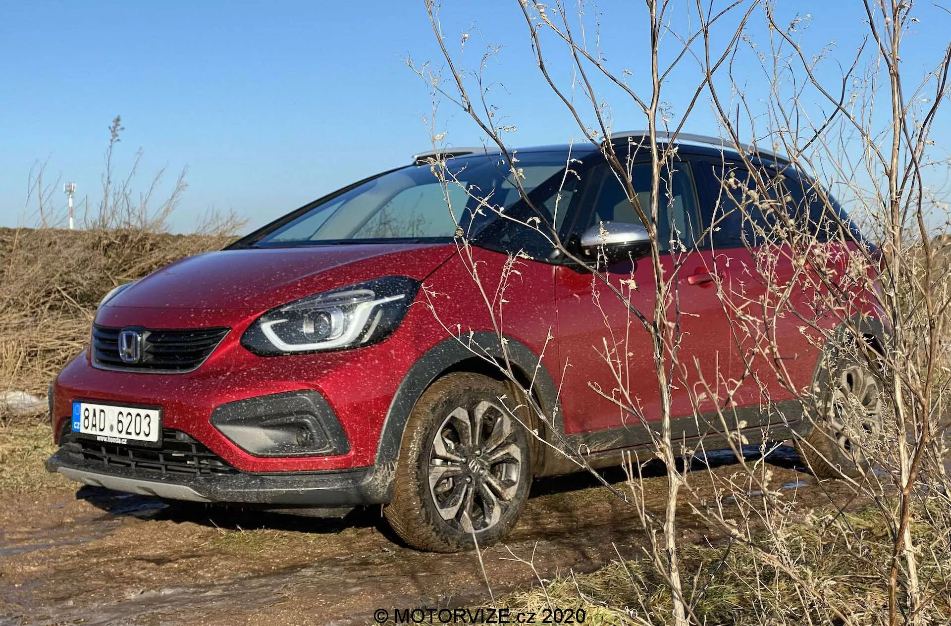 Foto diurna all'aperto di una Honda Jazz 2020 (Jazz Crosstar) parcheggiata su un terreno erboso. L'immagine cattura il colore rosso vibrante del SUV subcompatto, la griglia prominente e i moderni gruppi ottici con illuminazione a LED. La vettura è vista da un angolo frontale di tre quarti, mostrando l'altezza da terra aumentata, i cerchi in lega, i rivestimenti in plastica nera intorno ai passaruota e le parti inferiori della carrozzeria. Cielo azzurro chiaro, illuminazione naturale e rami circostanti oscurano parzialmente la sezione anteriore inferiore. Gli specchietti laterali con indicatori di svolta integrati e una vista dell'interno attraverso la finestra del conducente sono visibili.