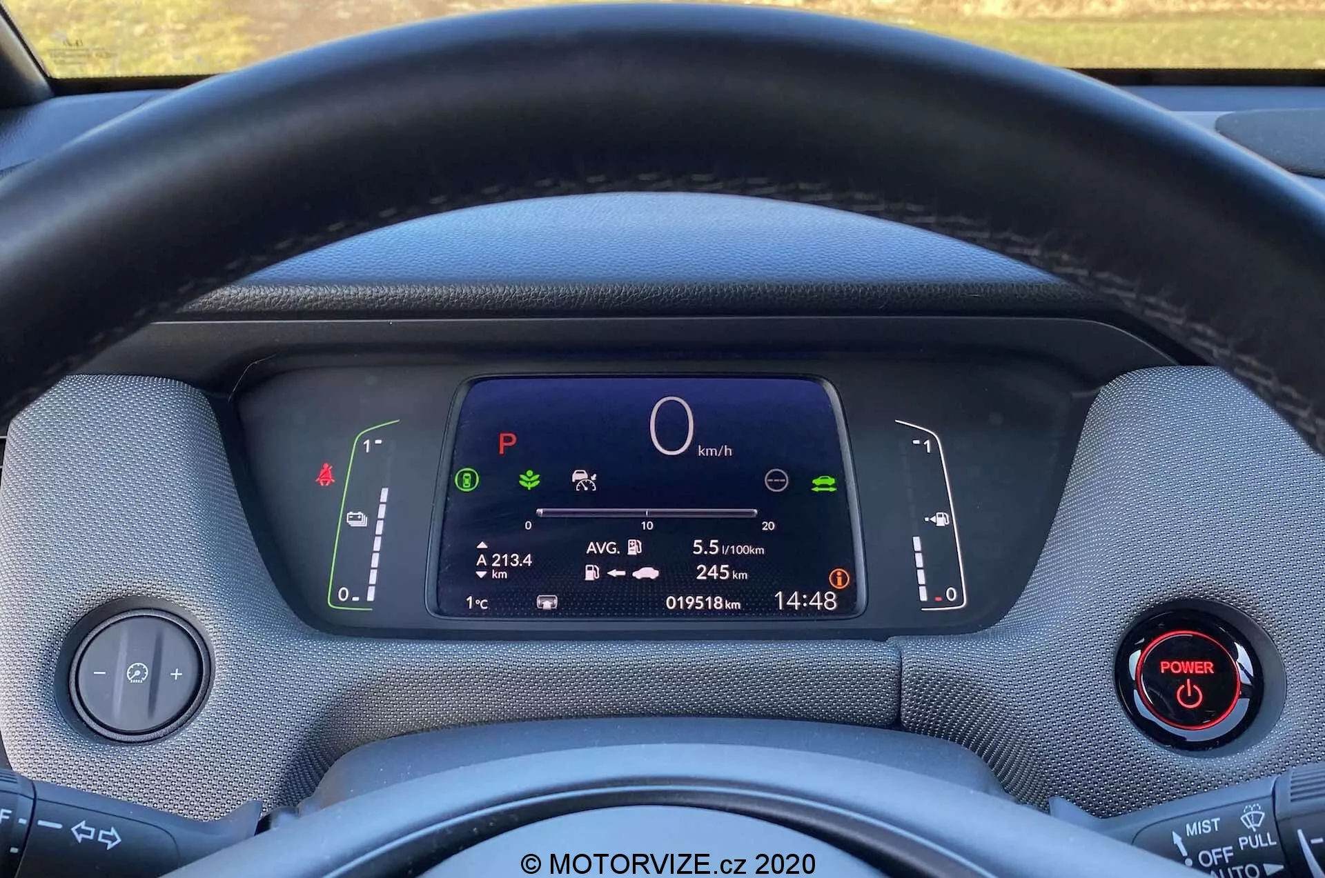 Primo piano degli interni della Honda Jazz Crosstar 2020 dal punto di vista del conducente, mostrando il volante e il quadro strumenti completamente digitale. Il volante rivestito in tessuto ostruisce parzialmente la vista del display digitale, che presenta simboli del sistema dell'auto, un grafico circolare centrale, informazioni sul viaggio, contachilometri e orologio. Due leve dietro il volante controllano gli indicatori di direzione, le luci e i tergicristalli. Un pannello di controllo per i fari e un pulsante di accensione con un anello rosso sono visibili rispettivamente a sinistra e a destra. I materiali del cruscotto sembrano strutturati, evidenziando un design ecologico. L'immagine enfatizza la tecnologia moderna, la strumentazione digitale e le potenziali caratteristiche ibride o elettriche del veicolo.