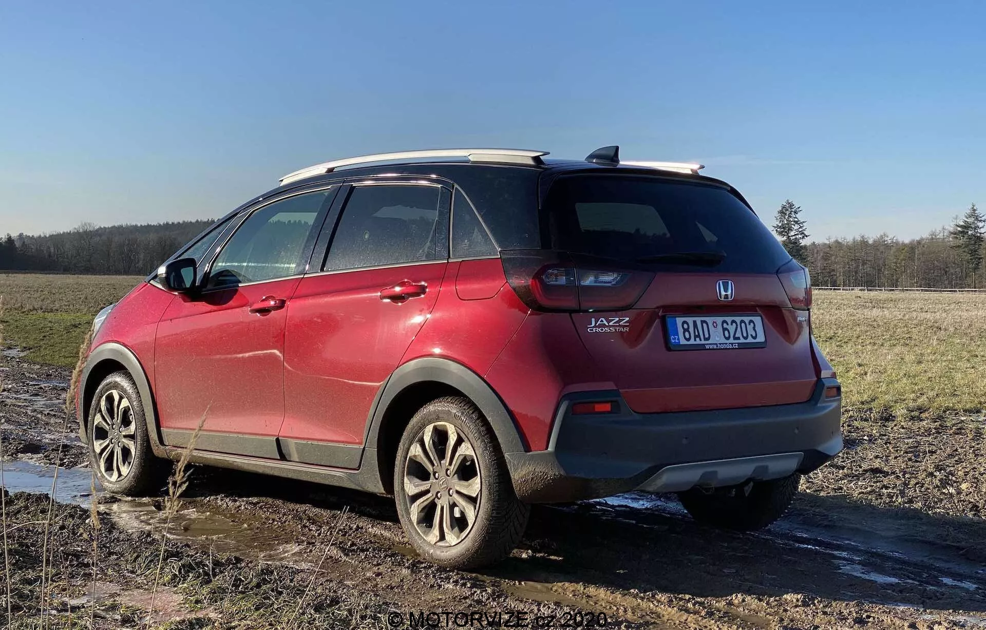 Vista posteriore di un quarto di un Honda Jazz Crosstar 2020 rosso, che mostra le caratteristiche del suo SUV crossover subcompatto. L'immagine catturata da un angolo basso evidenzia l'altezza e l'altezza da terra del veicolo, con gruppi ottici posteriori visibili, distintivo 'JAZZ', cerchi in lega, sistema di barre sul tetto, finestrini posteriori oscurati, schizzi di fango e una targa visibile, parcheggiato su un campo fangoso.