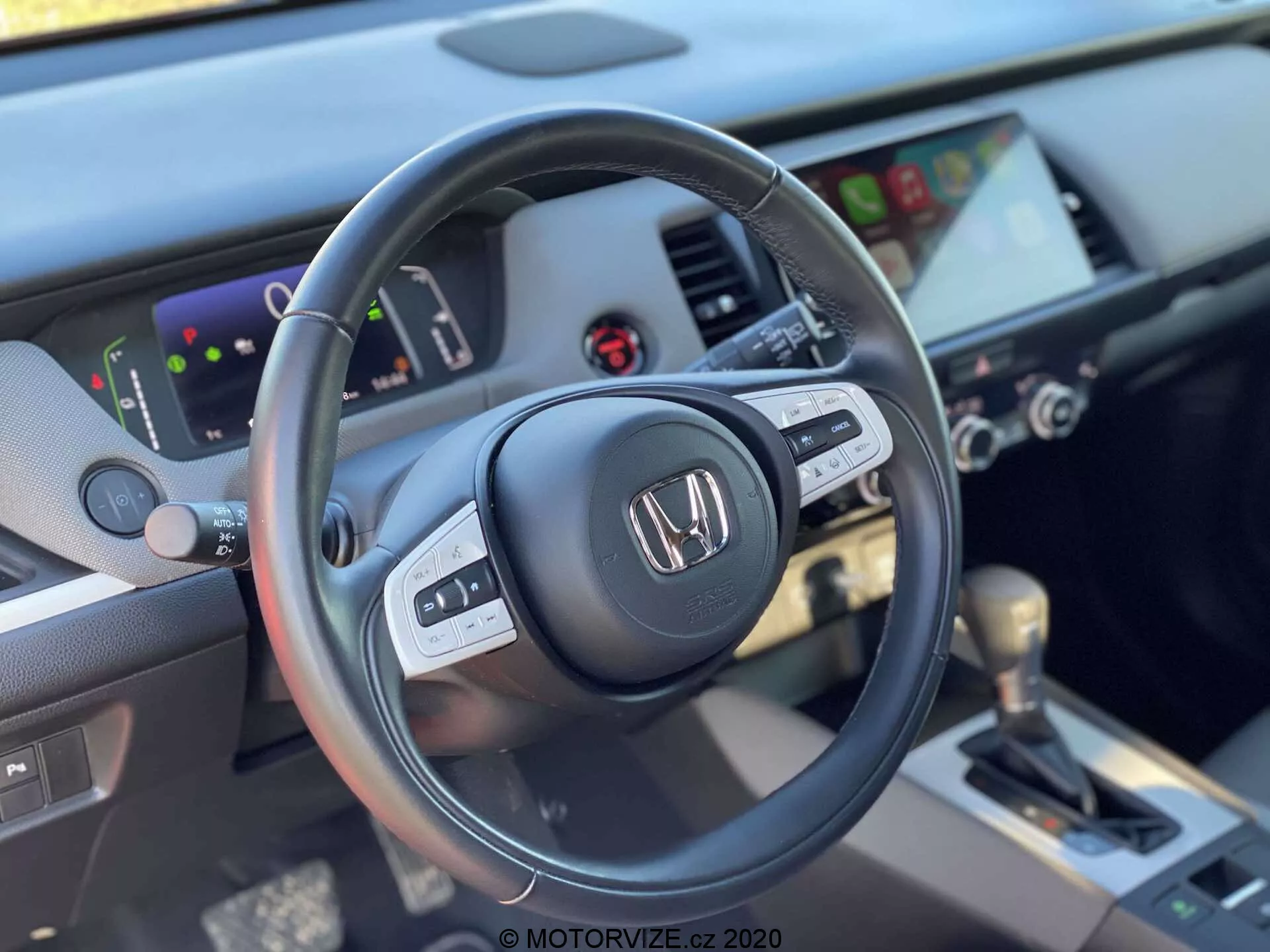 Vista ravvicinata dell'interno della Honda Jazz Crosstar 2020 presa dal sedile del conducente, che evidenzia il lato sinistro del volante a tre razze in pelle con comandi multifunzione e la plancia con un cruscotto digitale e un sistema di infotainment touchscreen. L'immagine include i comandi HVAC e una leva del cambio automatico con copertura in pelle nella console centrale, enfatizzando un design moderno e ben equipaggiato.
