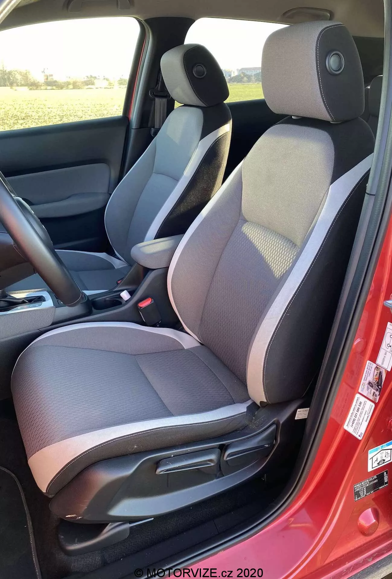 Vista interna della Honda Jazz Crosstar 2020 dal lato sinistro, che evidenzia il sedile del conducente in tessuto bicolore con sezione centrale a motivi, poggiatesta regolabile, controlli manuali di regolazione del sedile, cintura di sicurezza rossa, altoparlante sul pannello della porta, tappetino scuro, consolle centrale con bracciolo, controllo dello specchietto laterale e schema di colori scuro e chiaro.