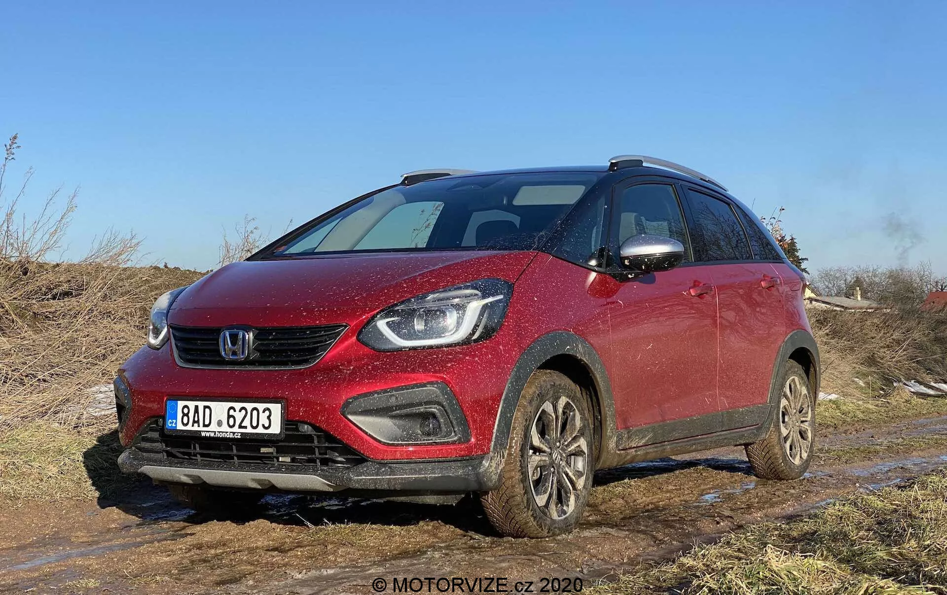 Vista angolare frontale di una Honda Jazz Crosstar 2020 rossa, un SUV crossover compatto, catturata su un terreno fuoristrada con terra e fango visibili. L'immagine mette in risalto la griglia anteriore, i fari eleganti che potrebbero ospitare lampade a LED o proiettore, i fendinebbia integrati nel paraurti e un cofano dolcemente inclinato. Il veicolo, dotato di barre sul tetto, ruote coperte di fango e specchietti laterali con indicatori di direzione integrati, suggerisce caratteristiche di sicurezza moderne e attrezzature tecnologiche avanzate. Ideale per categorie focalizzate su veicoli, recensioni di automobili, capacità fuoristrada, tecnologia automobilistica e design.