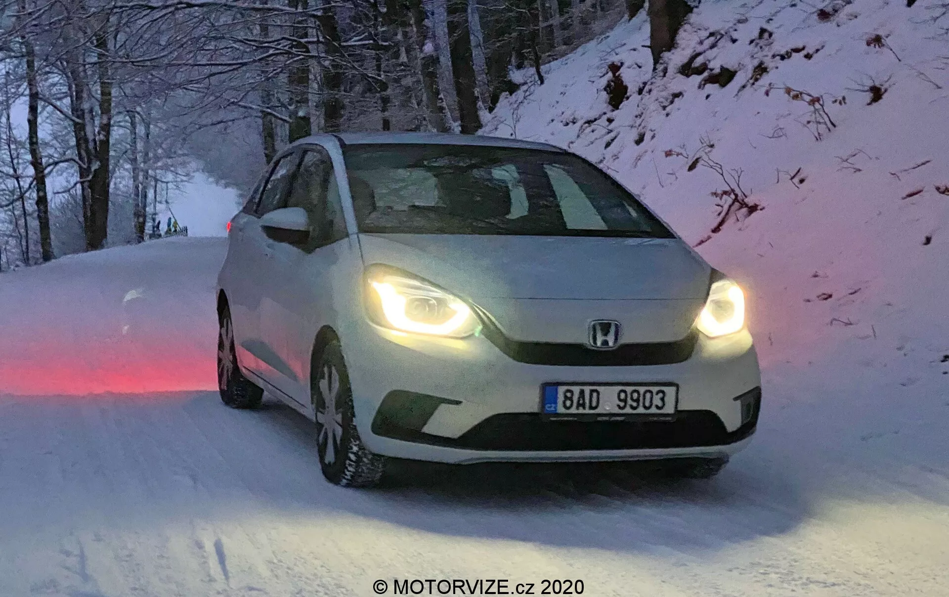 Questa immagine ritrae una Honda Jazz 2020, ripresa da un angolo anteriore di tre quarti, evidenziando le caratteristiche di design della griglia frontale, dei fari e dei contorni del cofano. Posizionata su una strada coperta di neve, il veicolo ha pneumatici invernali con battistrada visibile e i fari accesi, indicando condizioni di alba, tramonto o scarsa visibilità. La targa europea riporta “BAD 9903”, e l'immagine ha un leggero effetto di sfocatura che aggiunge una qualità dinamica. L'illuminazione naturale e fredda e l'ambientazione invernale sono completate dalle capacità del veicolo per tutte le stagioni.