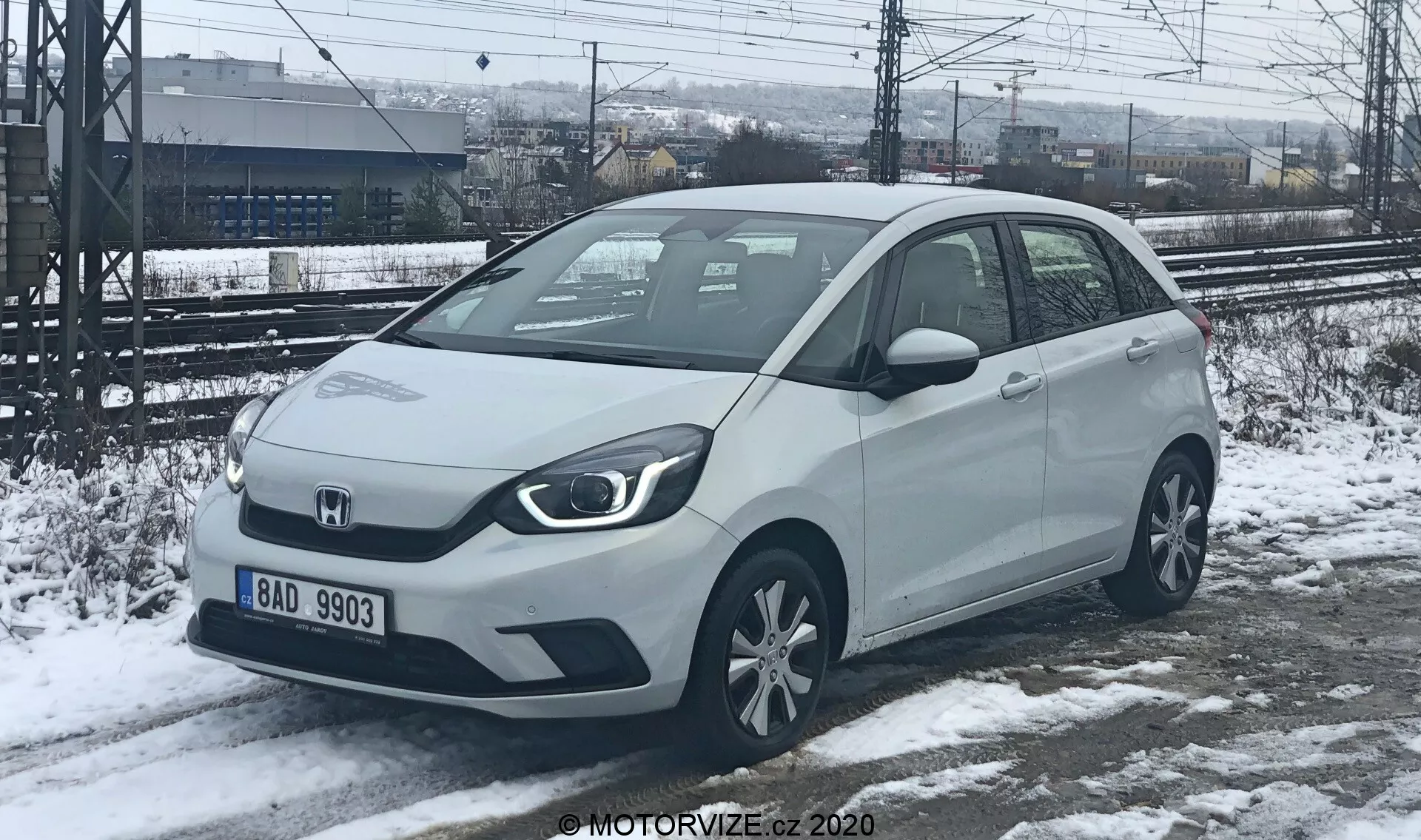 Vista angolare di tre quarti anteriore di un'auto compatta Honda Jazz 2020 in un colore chiaro, possibilmente bianco o argento, catturata in un contesto invernale con neve al suolo. I fari angolari e la griglia anteriore solida suggeriscono opzioni di motore elettrificato. L'auto presenta sensori di parcheggio visibili sul paraurti anteriore, specchietti laterali montati sulle porte con indicatori di direzione integrati e cerchi in lega standard. Parcheggiata su neve parzialmente sciolta, con binari ferroviari e una recinzione sullo sfondo, indicando una posizione industriale o vicino a una stazione ferroviaria.