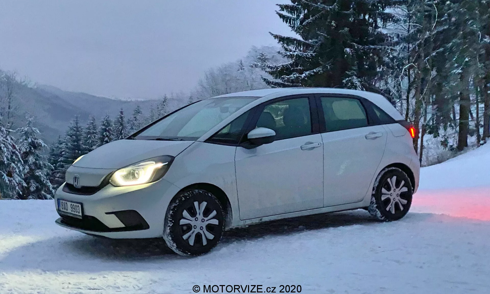 Honda Jazz 2020 hatchback compatta fotografata da un'angolazione frontale obliqua che cattura una vista completa del lato del conducente e una vista parziale del paraurti e della griglia anteriori. Il veicolo presenta uno schema di colore bicolore, con la maggior parte in una tonalità più chiara e la parte inferiore delle portiere laterali in una tonalità più scura. I fari sono accesi, con lampade alogene tipiche, senza fendinebbia. I copricerchi invernali decorativi sono visibili con fiocchi di neve riflettenti, indicando una stagione invernale. L'auto è parcheggiata su un'area coperta di neve con uno sfondo di alberi e una collina innevata, suggerendo una posizione rurale o montuosa. Immagine per gentile concessione di MOTORVIZE.cz.