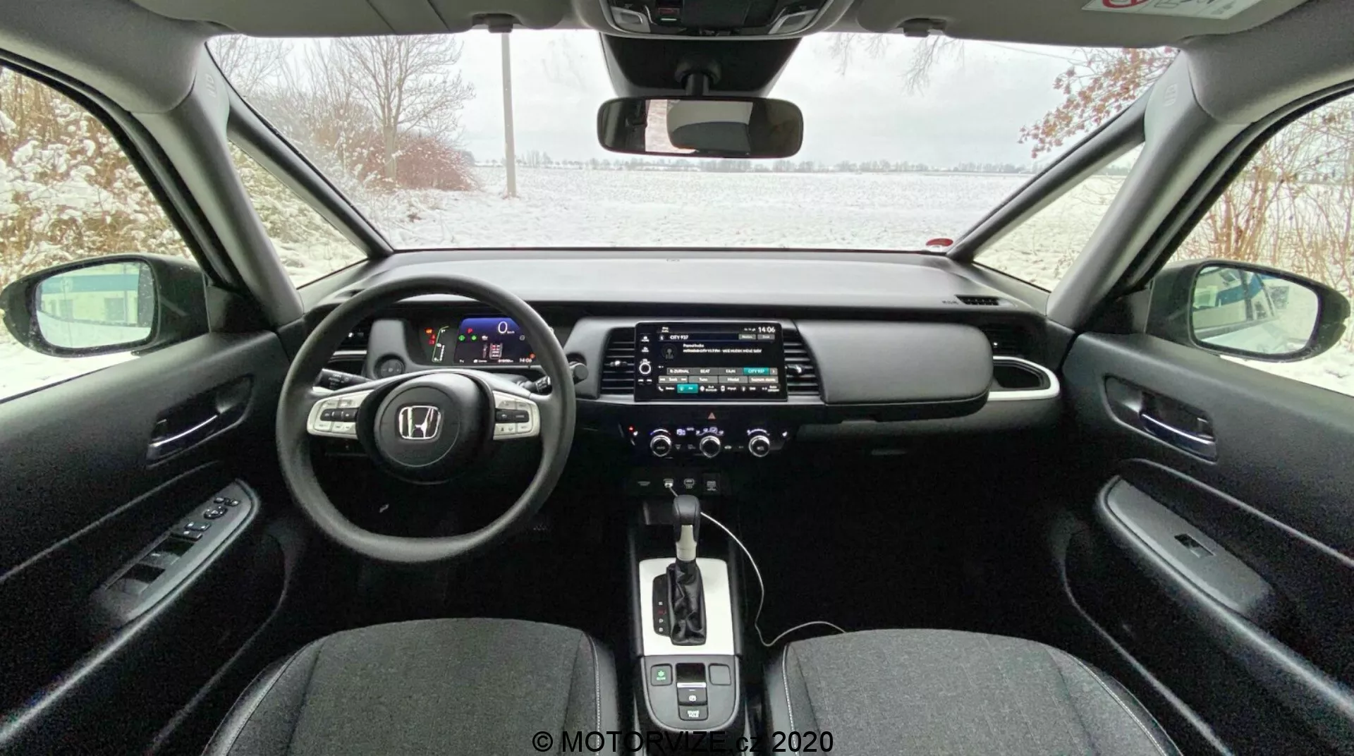 Interni di un'auto moderna visti dal sedile posteriore, con quadro strumenti digitale, volante multifunzionale, grande touchscreen infotainment, cambio automatico con pomello in pelle, climatizzatore bizona, freno di stazionamento elettronico e design elegante con materiali a contrasto; esterno innevato visibile dal parabrezza, che suggerisce una possibile ambientazione invernale.