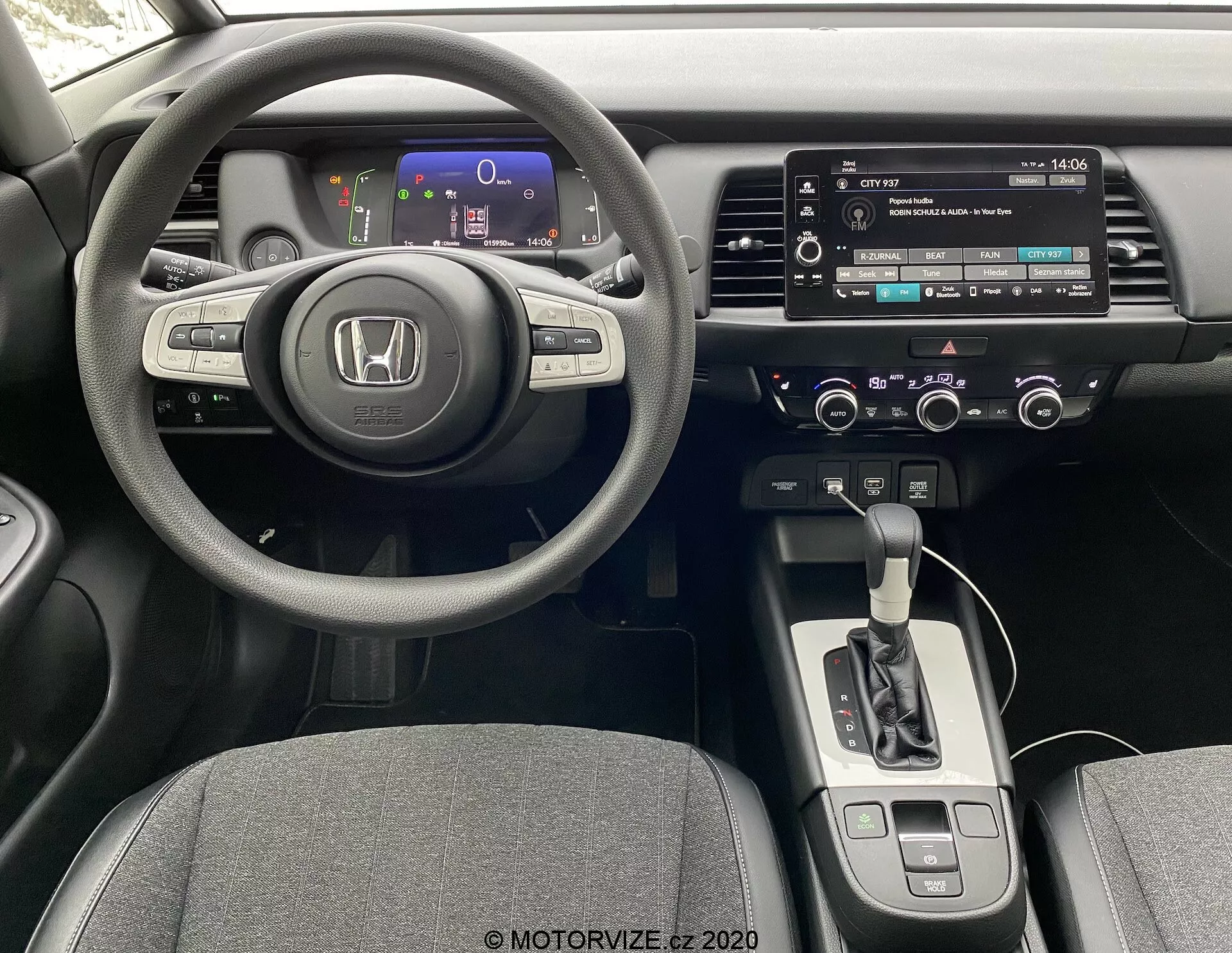 Interni di una Honda Jazz 2020 dalla prospettiva del conducente, con un sofisticato volante a tre razze con comandi integrati, un quadro strumenti digitale con indicatori colorati, una console centrale con sistema di infotainment touchscreen, controlli manuali per audio e climatizzazione, un selettore del cambio automatico, e sedili rivestiti in tessuto.