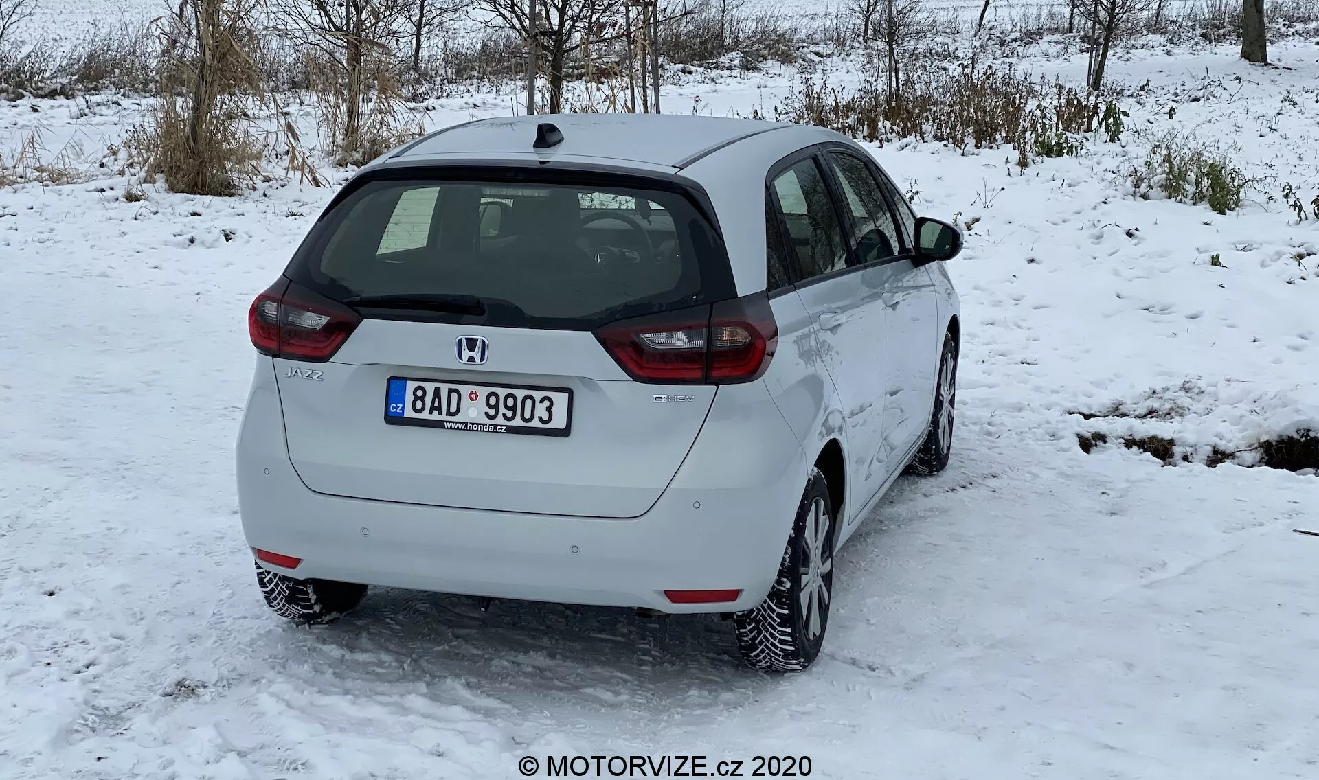 L'immagine mostra una vista posteriore tre quarti di una Honda Jazz 2020 bianca, parcheggiata su una superficie coperta di neve, con alberi senza foglie sullo sfondo invernale e un cielo nuvoloso. Le caratteristiche del design posteriore includono un tergicristallo posteriore, il logo del produttore, l'indicazione del modello e l'assemblaggio dell'illuminazione posteriore con lenti rosse e chiare. Il profilo laterale mostra la porta passeggeri posteriore, la porta anteriore, i finestrini laterali e un tappo del carburante visibile. L'auto è dotata di cerchi in lega a più razze, con battistrada adatto alla neve e una leggera accumulazione di neve. La targa posteriore suggerisce la sua origine geografica. L'altezza da terra è tipica delle hatchback, senza terminale di scarico visibile né accessori esterni. L'auto sembra pulita e in buone condizioni, con poca sporcizia sul paraurti posteriore dovuta a strade innevate.
